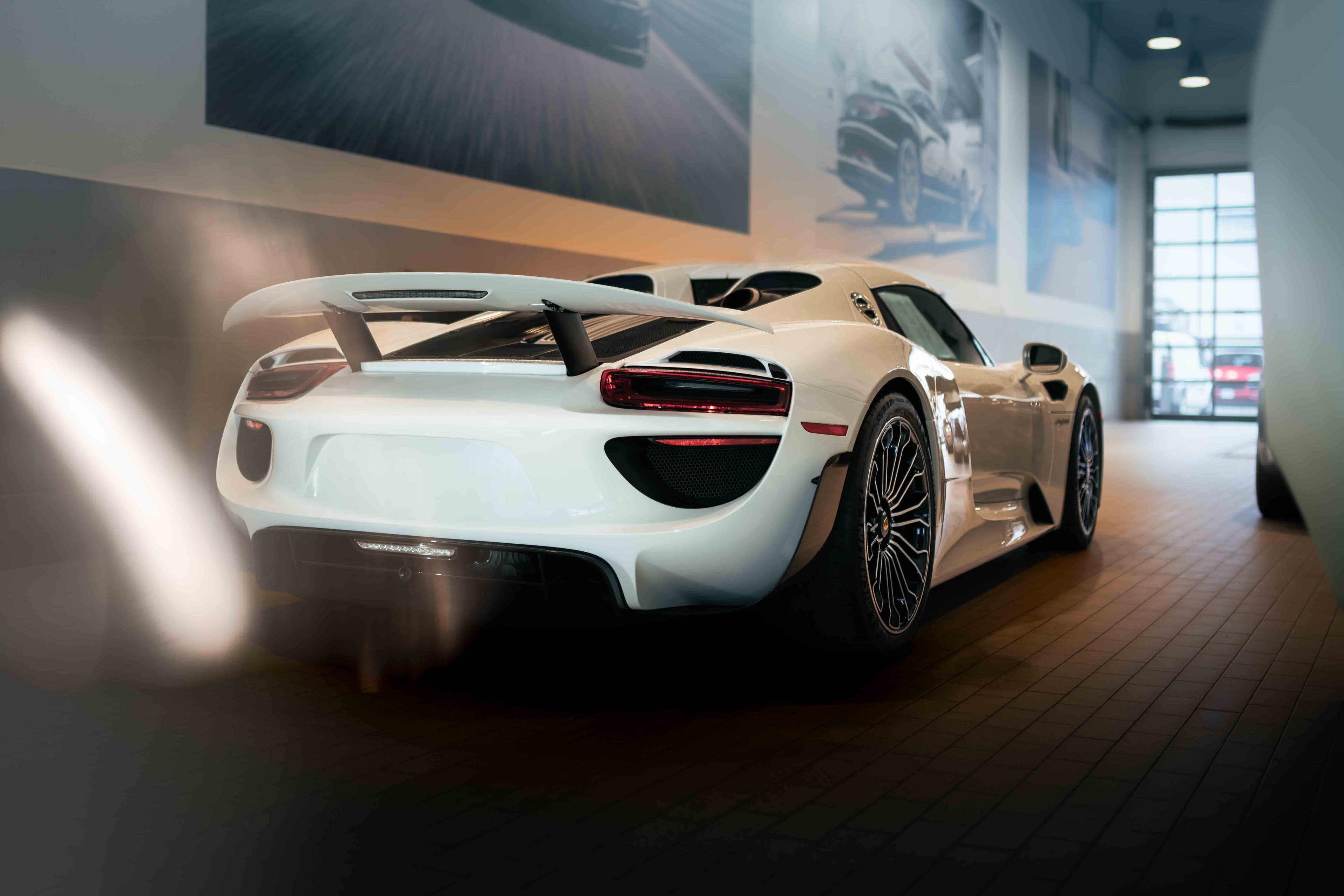 White Porsche 918 Spyder emitting exhaust vapour inside a high-end dealership, with dynamic reflections and automotive posters on the wall.