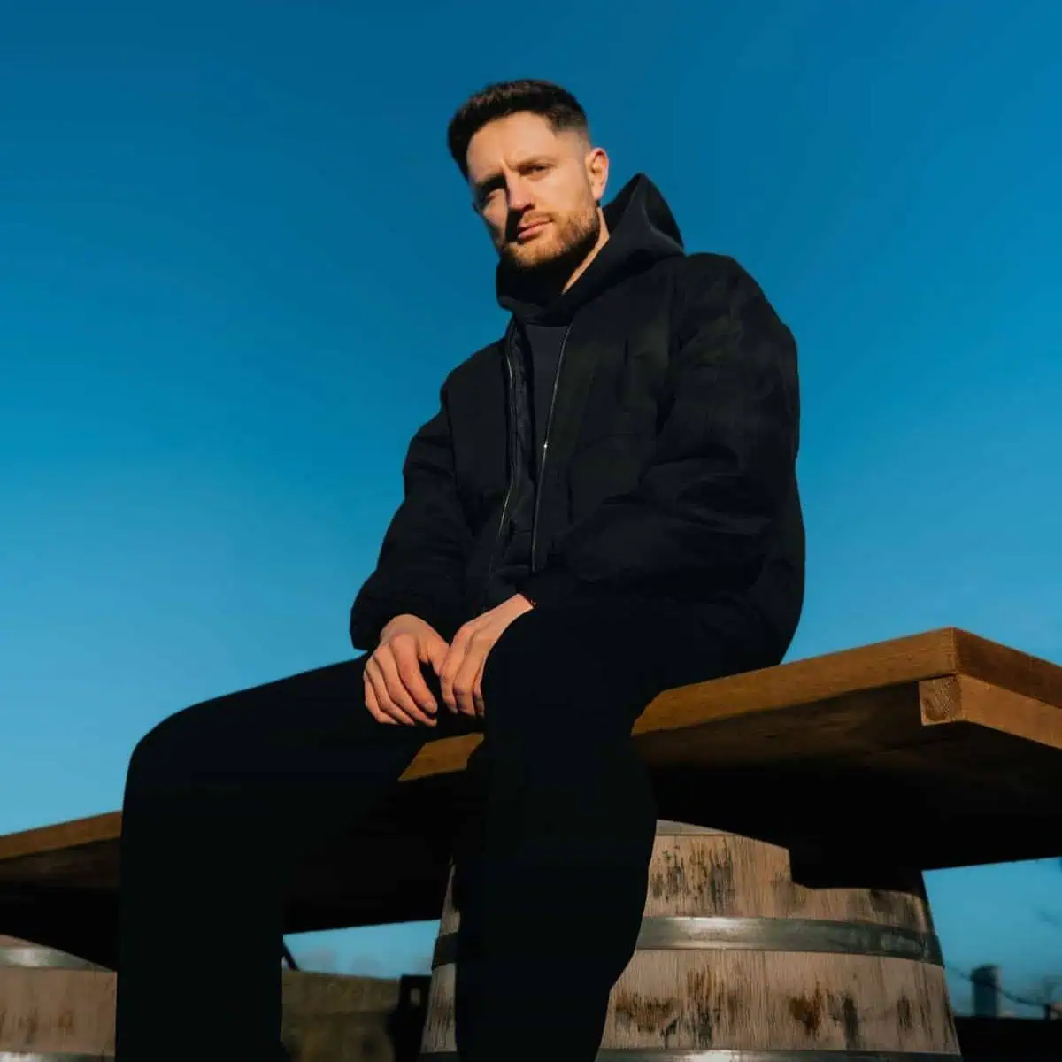 Simon Cousins, founder of TIRES + TERRAIN and motorsport engineer, seated on a wooden bench supported by barrels, wearing a black jacket against a vibrant blue sky backdrop.