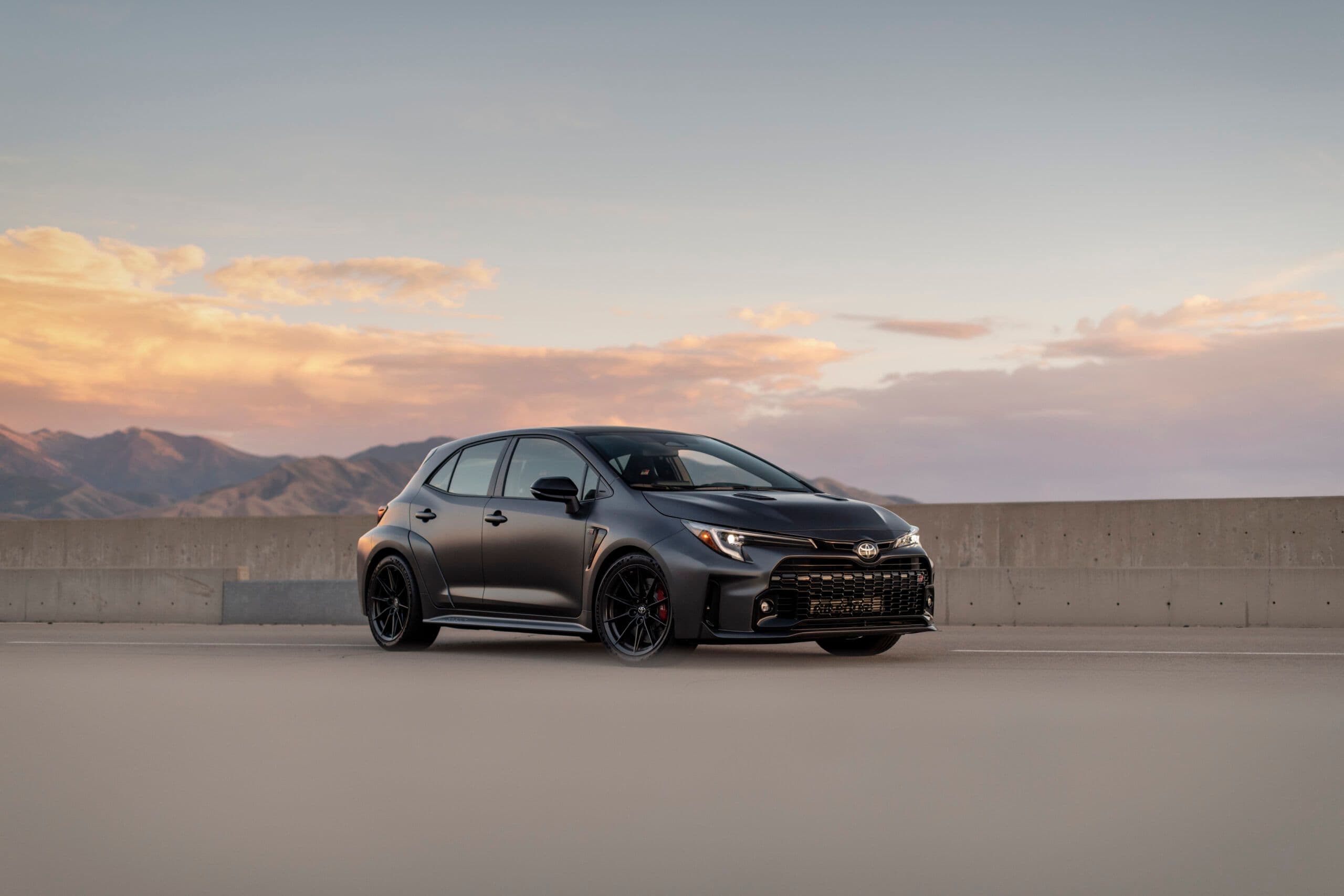 A matte black Toyota GR Corolla parked on a rooftop with a mountain range and soft pastel sunset sky in the background.