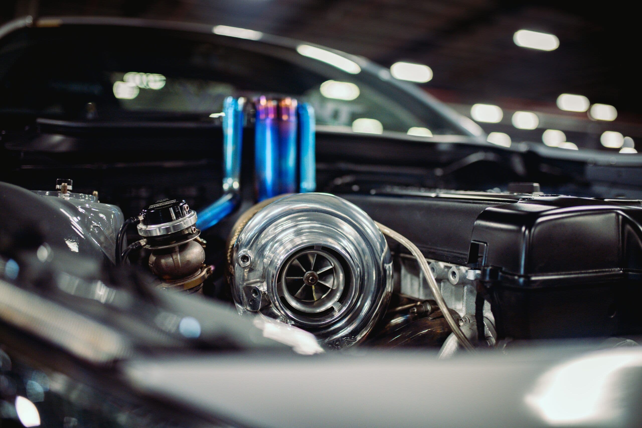 A high-performance turbocharger setup inside an engine bay, featuring polished metal housing and heat-treated titanium piping, with a blurred garage background.