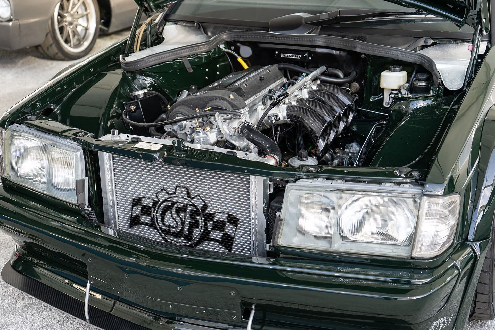 Engine bay of a modified Mercedes-Benz 190E featuring a high-performance CSF radiator and individual throttle bodies, showcasing a clean and detailed build with a dark green paint finish.
