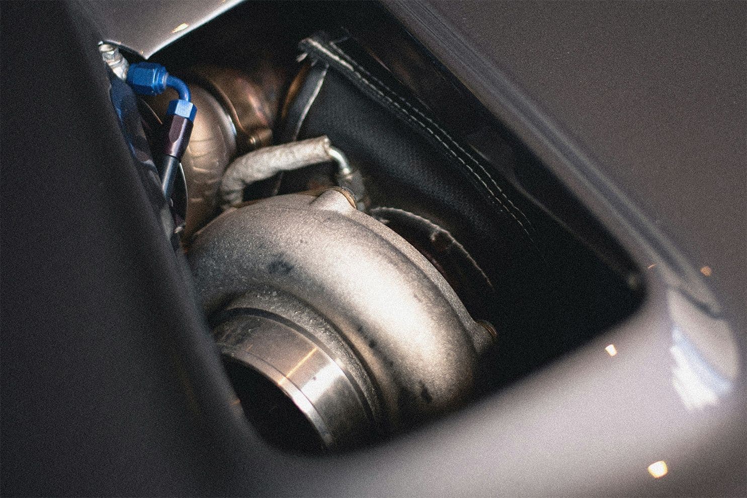 Close-up of a turbocharger visible through the bonnet vent of a performance car, showing heat shielding and braided fuel lines.