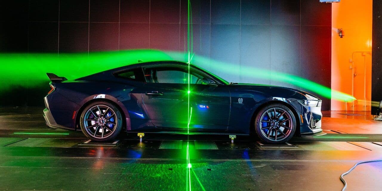 A side profile of a dark blue Ford Mustang Dark Horse undergoing aerodynamic testing in a wind tunnel, with green airflow visualisation and laser scanning beams highlighting drag pathways.