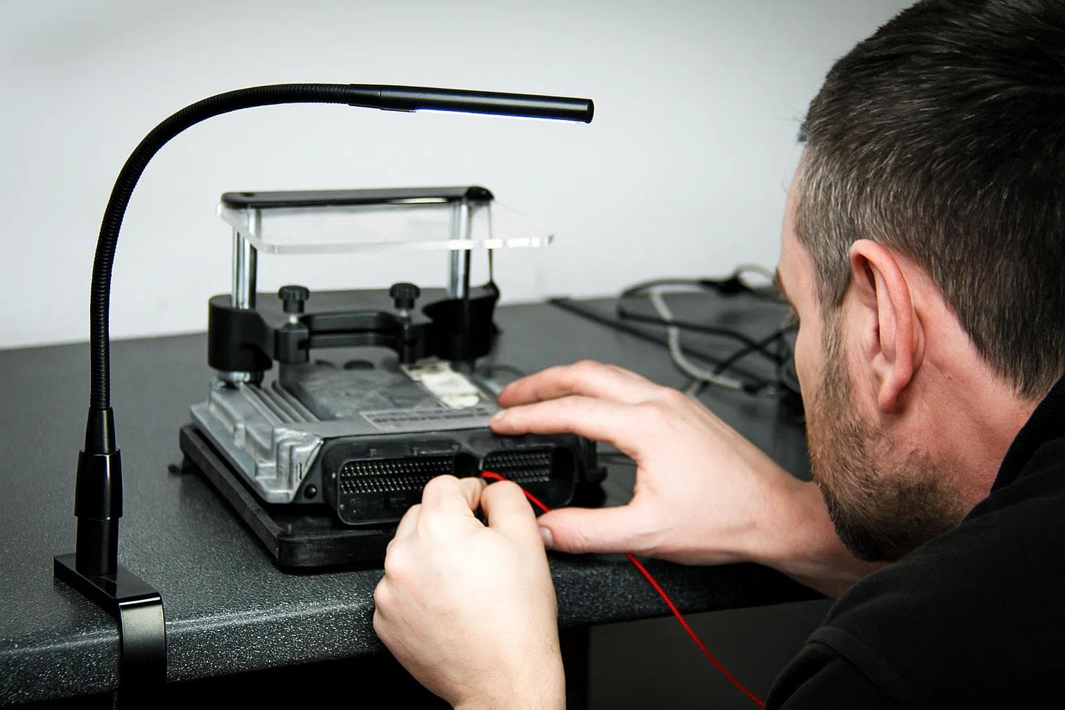 Engine tuner connecting wiring to an ECU on a bench setup for programming or remapping, under a flexible desk light, with a focused and precise setup.