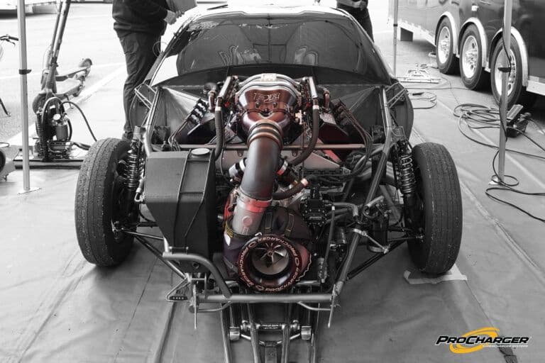 Rear view of a stripped-down drag car chassis showcasing a ProCharger centrifugal supercharger setup with exposed piping, suspension, and engine components in a race paddock.