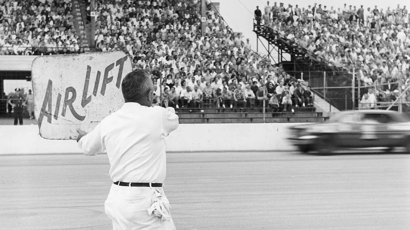 Historic motorsport image showing a pit crew member holding an “Air Lift” sign at a race