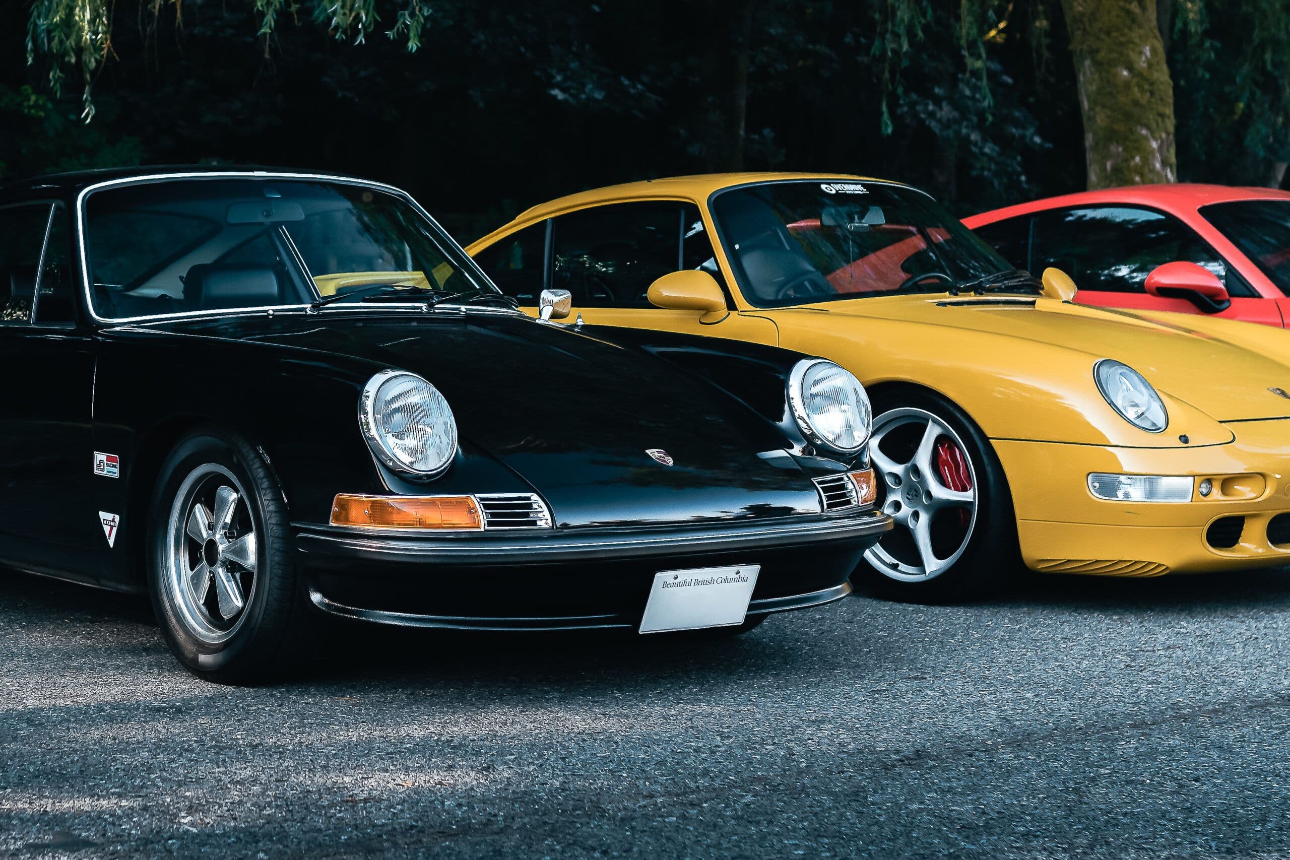 A row of classic and modern Porsche 911 models parked outdoors, featuring a black long-hood 911, a yellow 993, and a red 996, with trees in the background.
