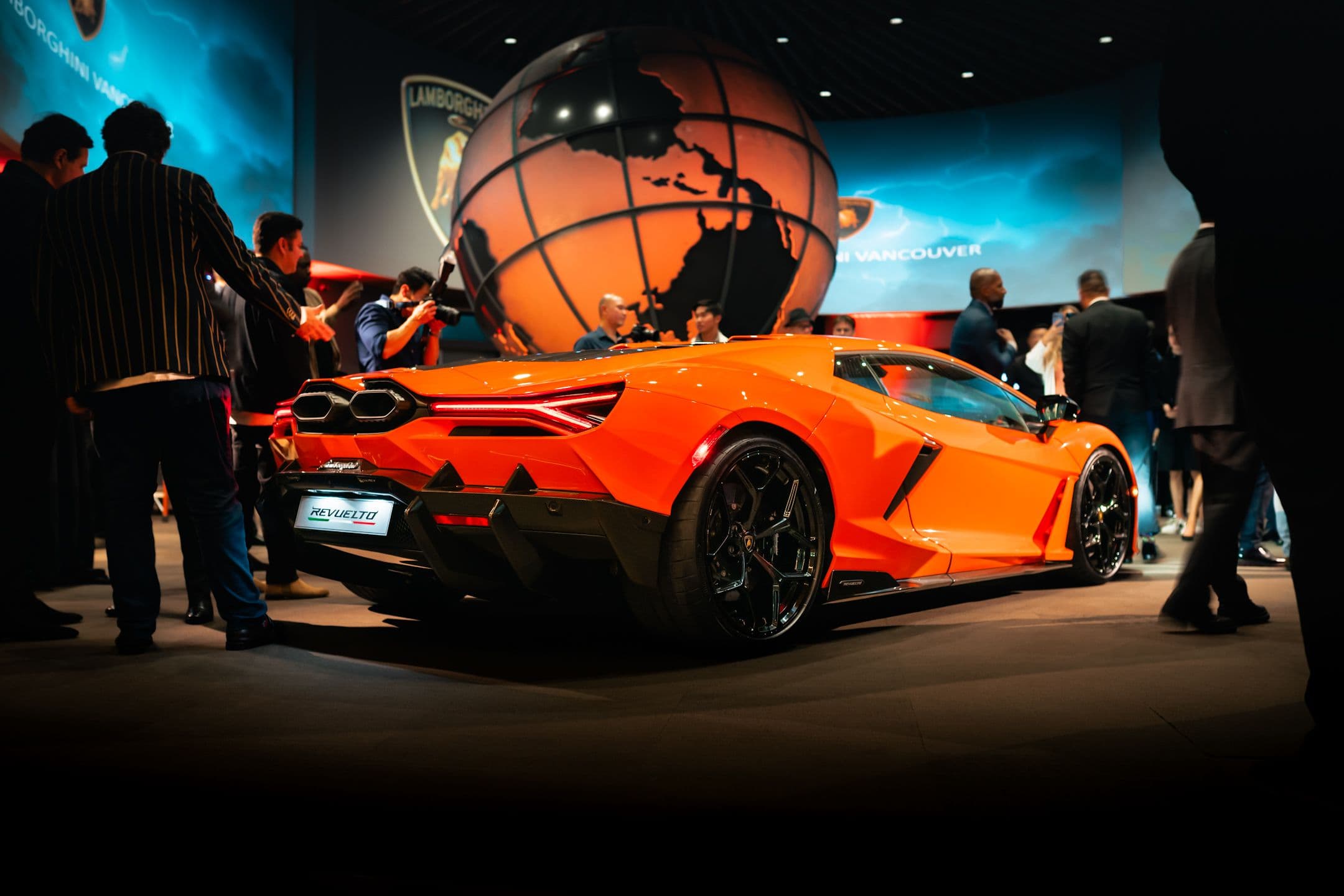 Lamborghini Revuelto in Arancio Apodis at Vancouver launch event, rear three-quarter angle with crowd and globe display.
