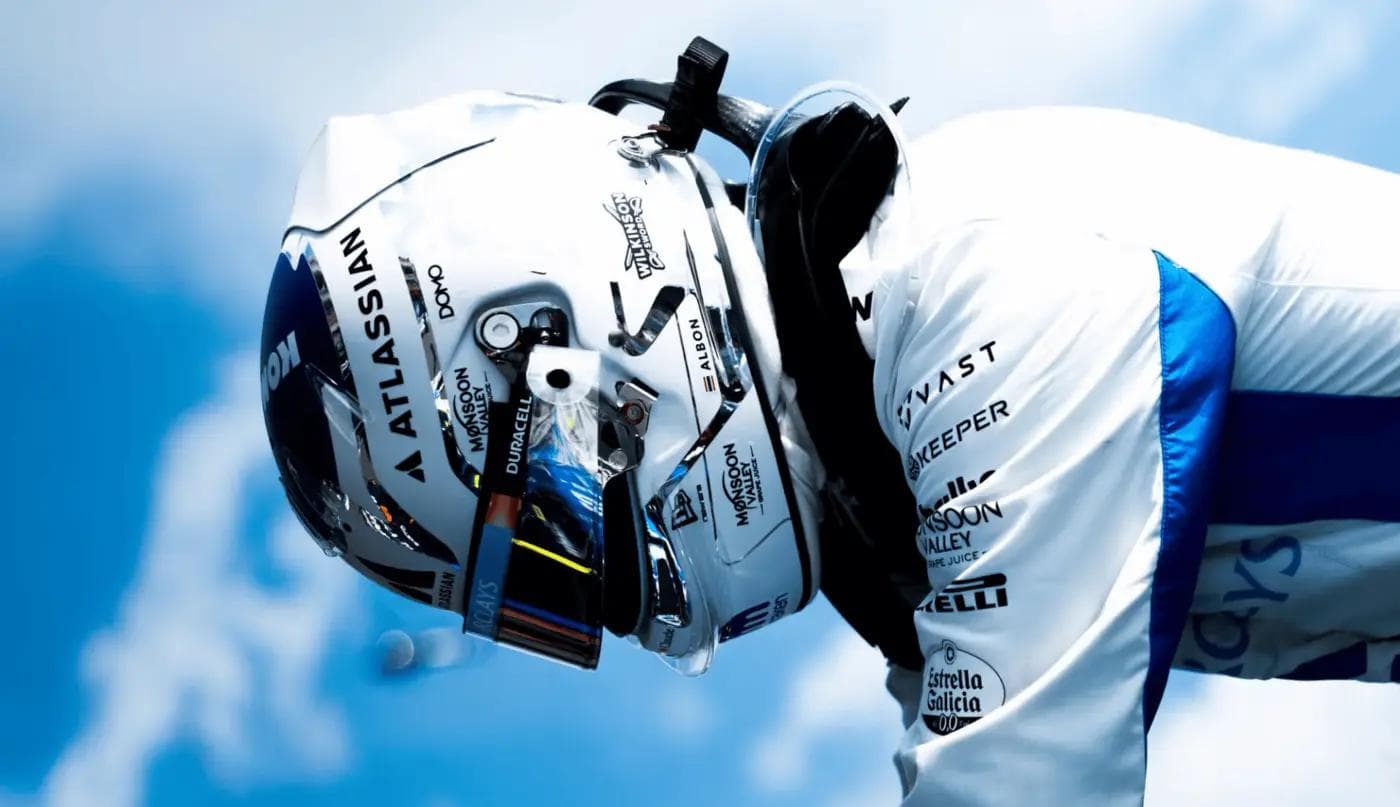 Williams Racing Formula 1 driver Alex Albon in white helmet and race suit against a blue sky, sponsor logos including Atlassian and Duracell visible on livery