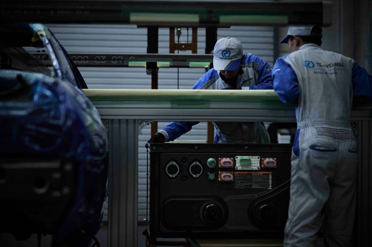 Nissan engineers in branded uniforms assembling components of the Nissan Skyline R32 GT-R EV in a workshop. The image highlights the meticulous craftsmanship involved in converting the iconic car to electric power.
