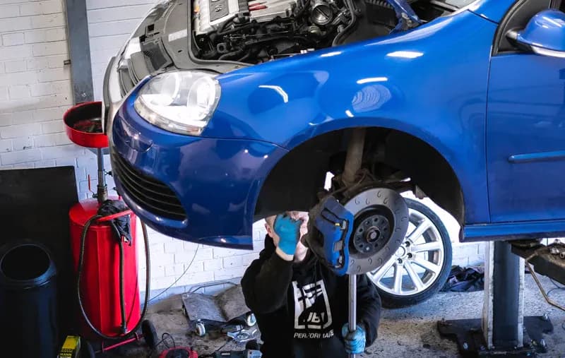 Technician performing a pre tuning inspection on a blue Volkswagen Golf Mk5 on a workshop lift, checking suspension and brake components with the front wheel removed.