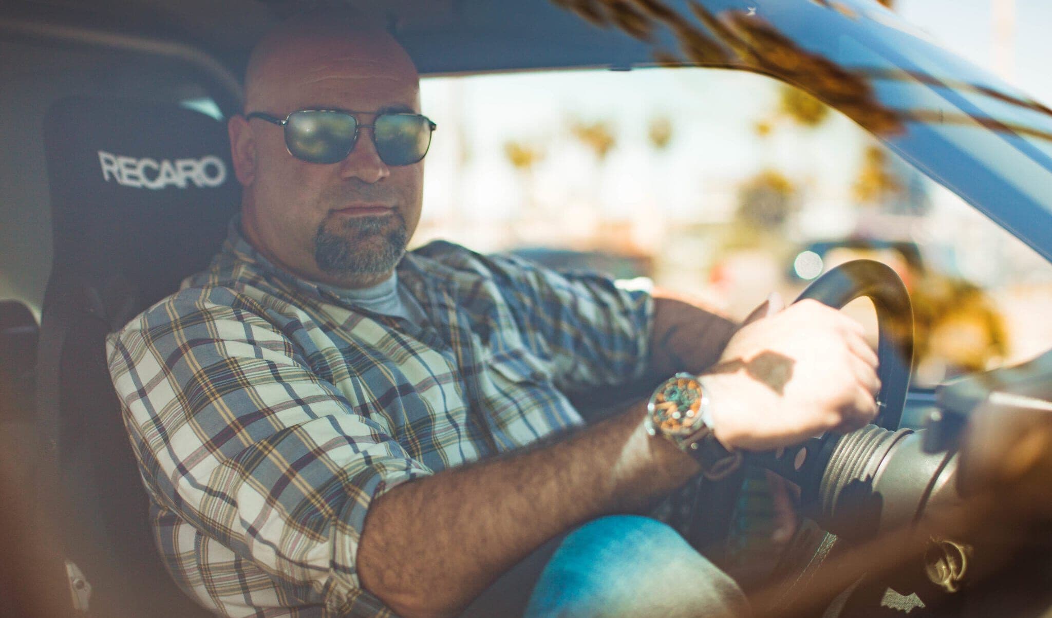 Matt Farah of The Smoking Tire seated in a car with a Recaro racing seat, wearing sunglasses, a plaid shirt, and a prominent wristwatch, photographed from outside the driver-side window on a sunny day.