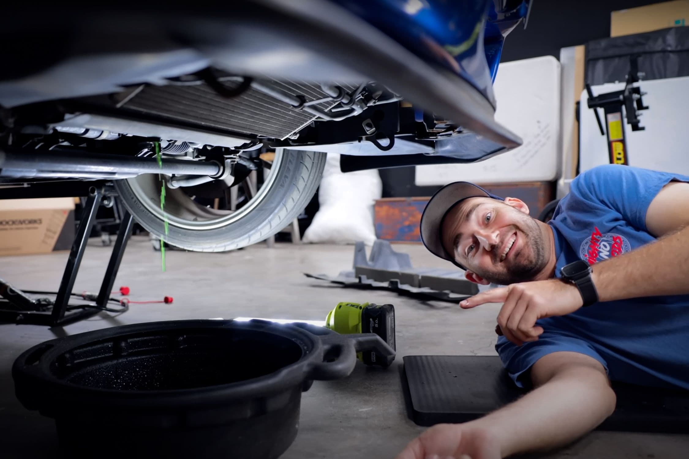 Marty from Mighty Car Mods lying on the garage floor, smiling and pointing under a blue car while coolant drains into a black catch pan during a DIY service.