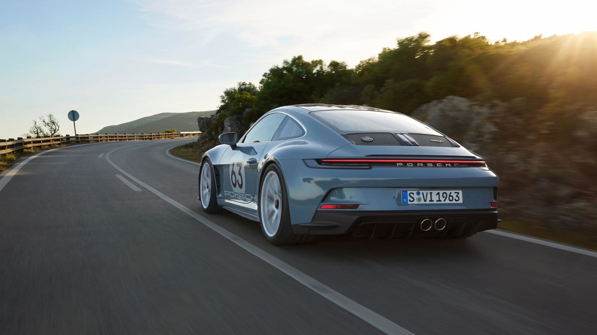 Rear view of blue Porsche 911 ST 992 driving on mountain road at sunset, wearing number 63 livery.