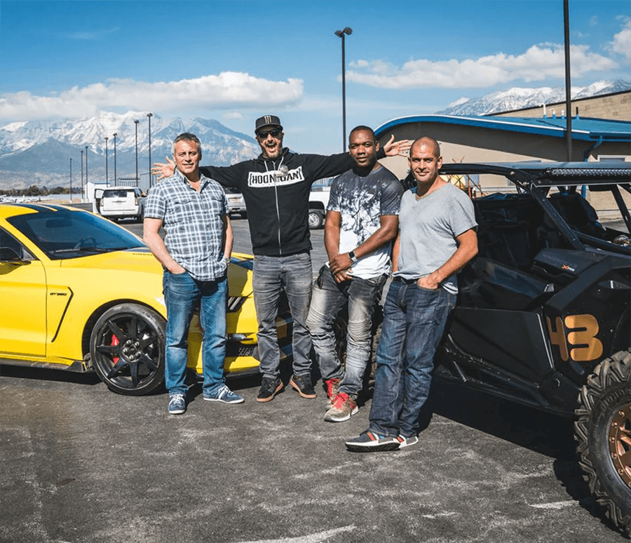 Ken Block posing with Top Gear hosts Chris Harris, Matt LeBlanc, and Rory Reid, standing between a yellow Ford Mustang and an off-road buggy, with mountains and a clear blue sky in the background.