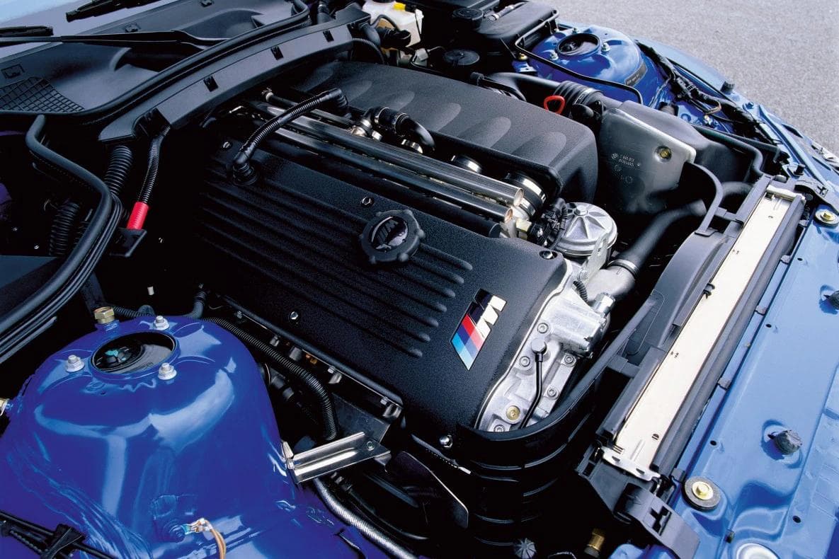 Close-up view of a BMW Z3 M Coupe engine bay featuring the high-revving S54 inline-six engine with the iconic M Power logo on the valve cover, finished in a metallic blue chassis.
