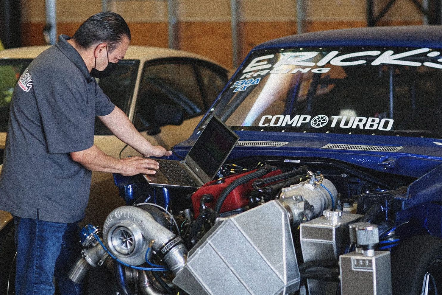 Tuner calibrating ECU on a Honda K-series drag car with a large single turbo, front-mount intercooler, and COMP Turbo hardware during engine tuning session.