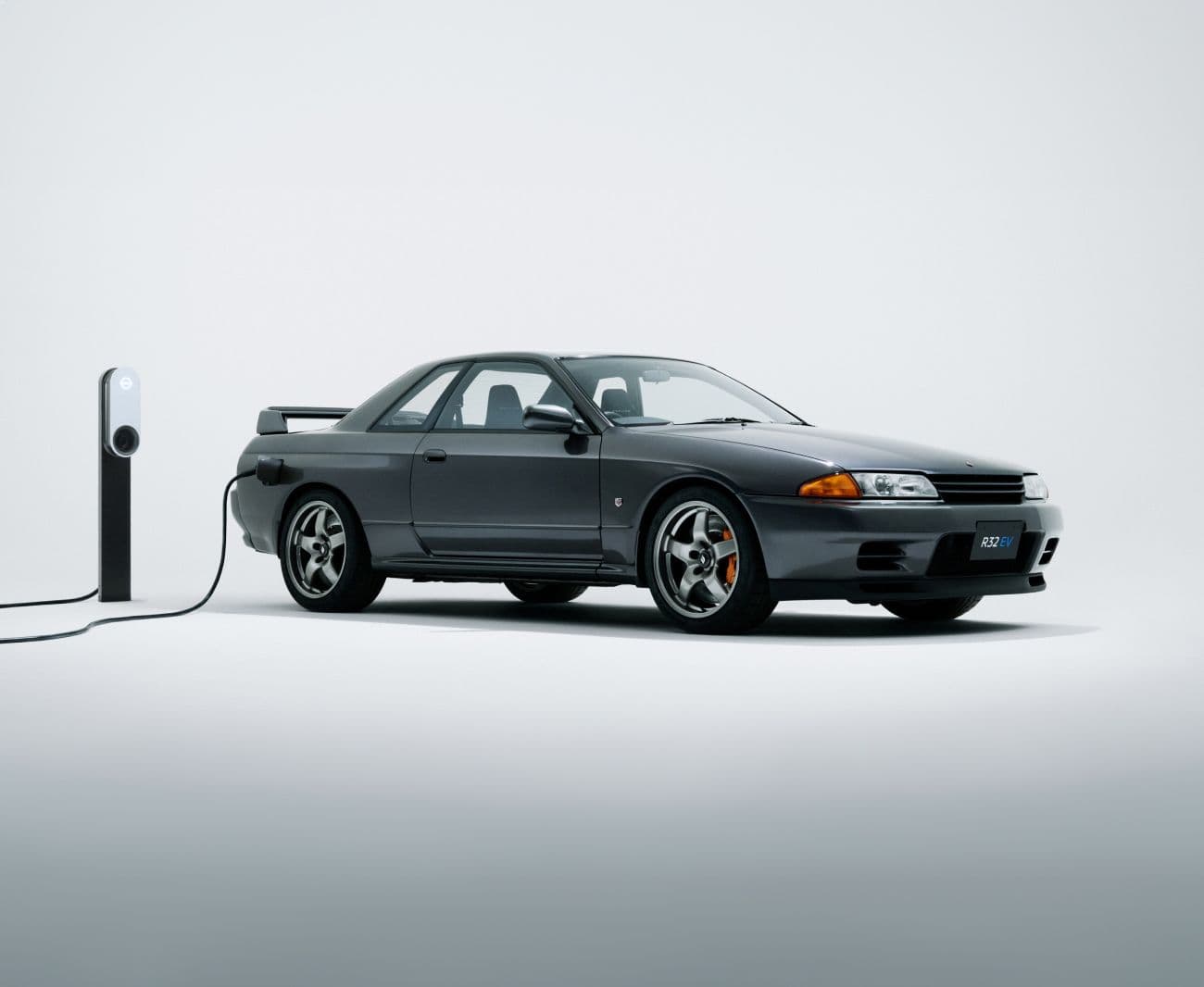 The Nissan Skyline R32 GT-R EV, a retrofitted electric version of the iconic sports car, plugged into a charging station. Sleek and modernized for zero-emissions performance.