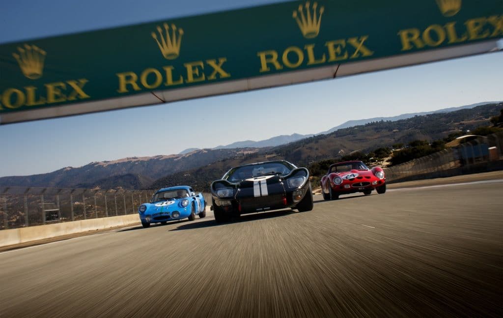Three vintage race cars – a blue Shelby Daytona Coupe, a black Ford GT40, and a red Ferrari 250 GTO – drive side by side under a Rolex banner on a scenic racetrack with mountains in the background.