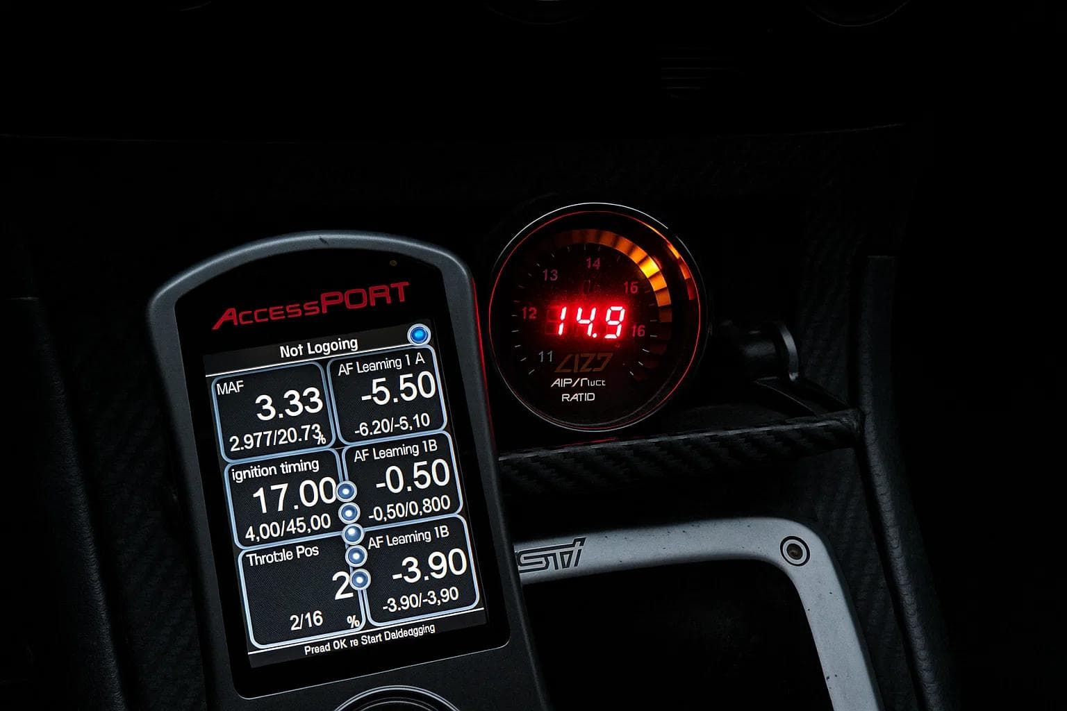 Close-up of a Cobb AccessPORT handheld tuner displaying live engine data alongside an AEM digital air-fuel ratio gauge reading 14.9 in a Subaru STI interior.