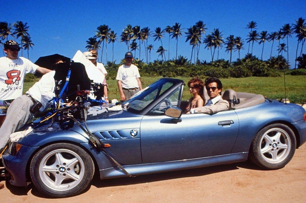 Behind-the-scenes photo of a film crew shooting a scene from a James Bond movie featuring a blue BMW Z3 Roadster with actors seated inside, surrounded by palm trees and tropical scenery.