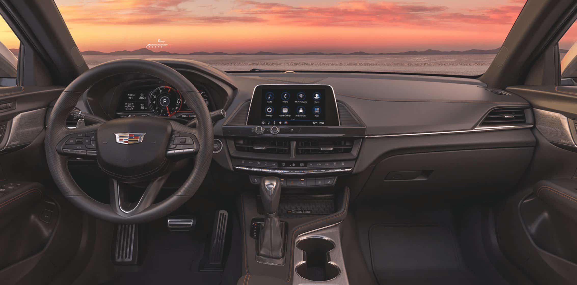 Interior view of a Cadillac with a sunset landscape through the windshield, showcasing the digital cluster, infotainment screen, and stitched leather dashboard.