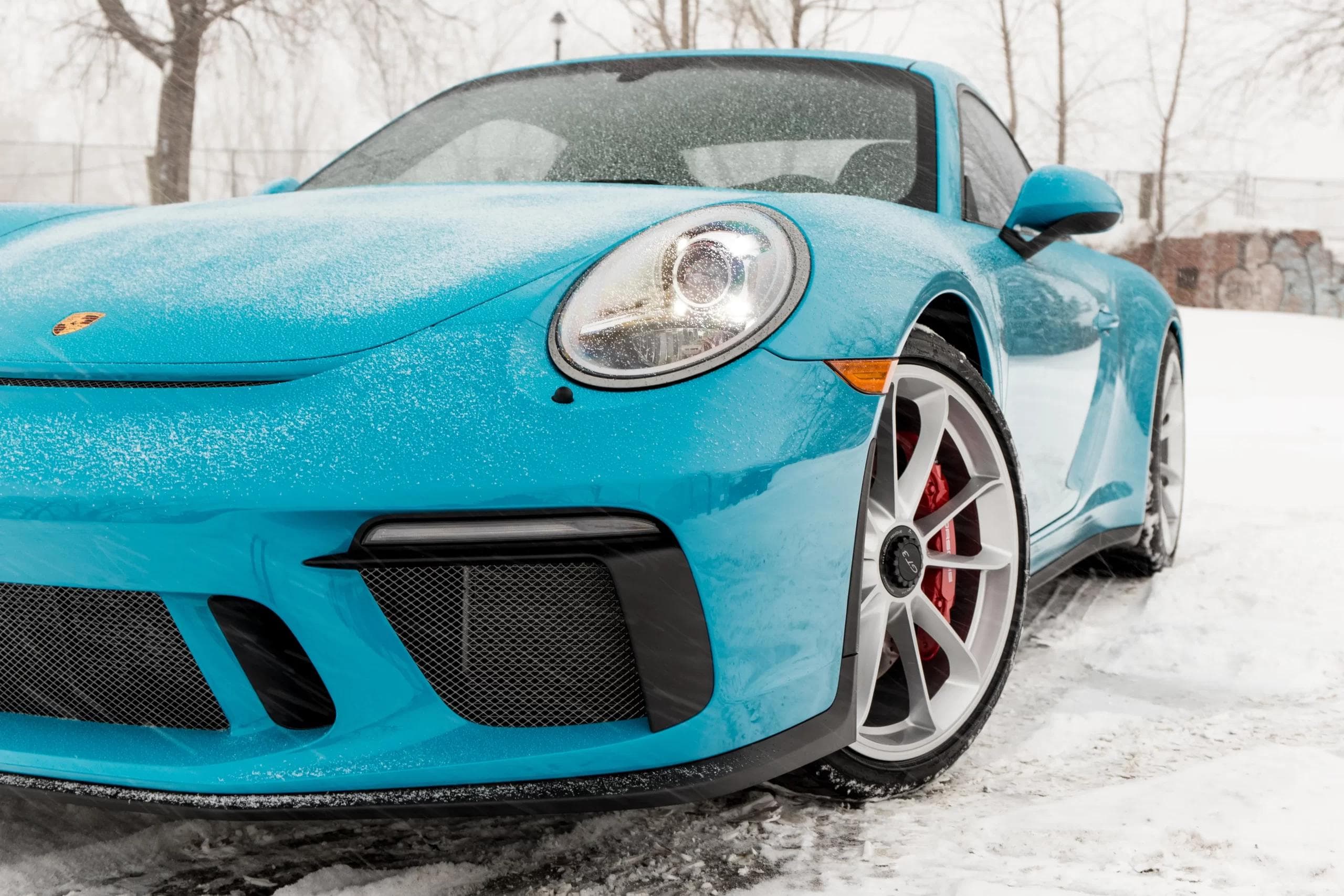 Front angle of a bright blue Porsche 911 Carrera with snow dusted over the bonnet and wheels, parked in a wintry urban setting.