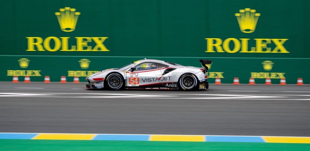 A Ferrari 458 GTE race car with number 54 livery and VistaJet sponsorship speeding along the Rolex-branded barriers at the 24 Hours of Le Mans circuit.