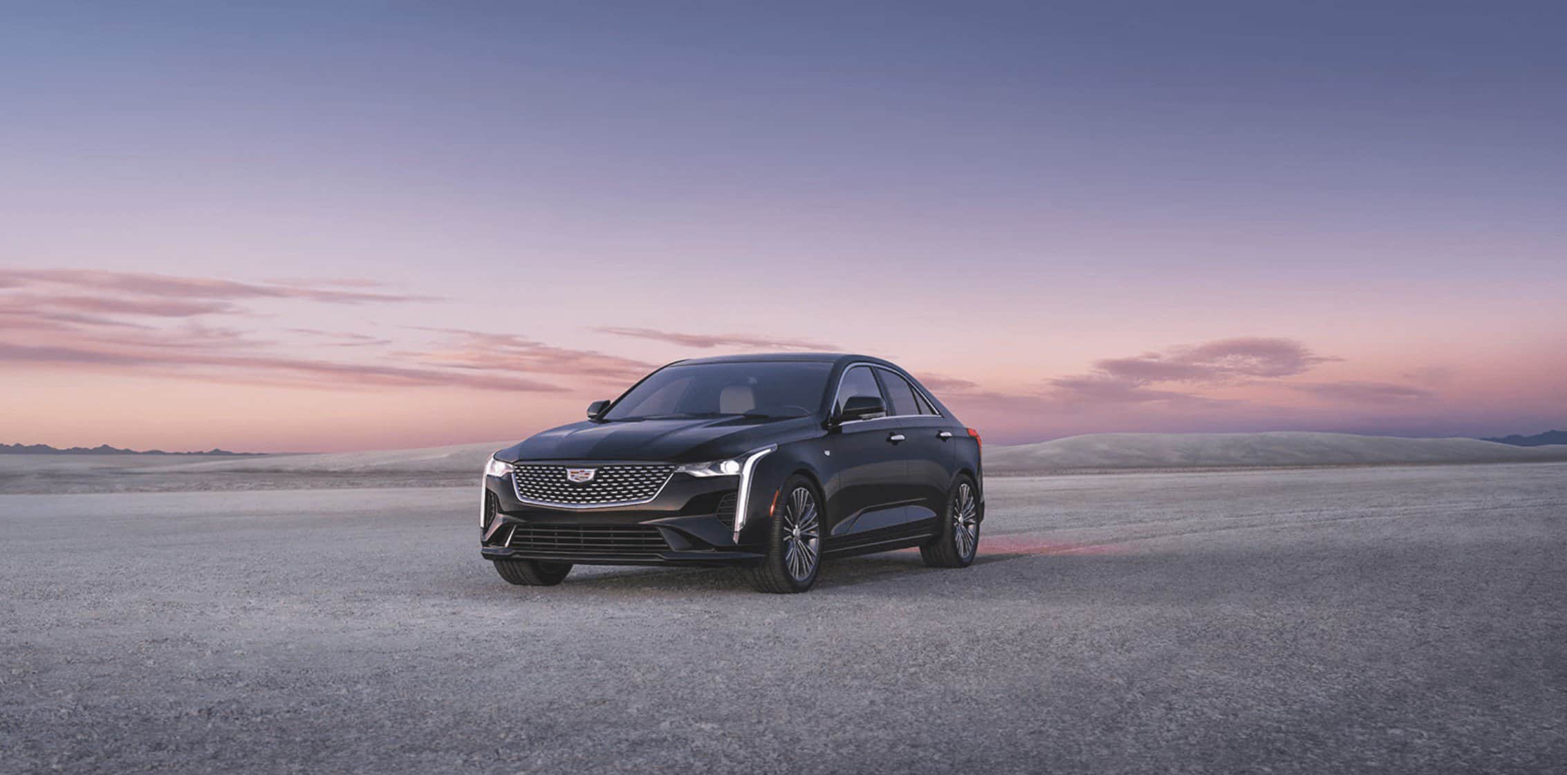 A black Cadillac CT4 sedan parked on a vast desert plain under a soft purple and pink sunset sky.