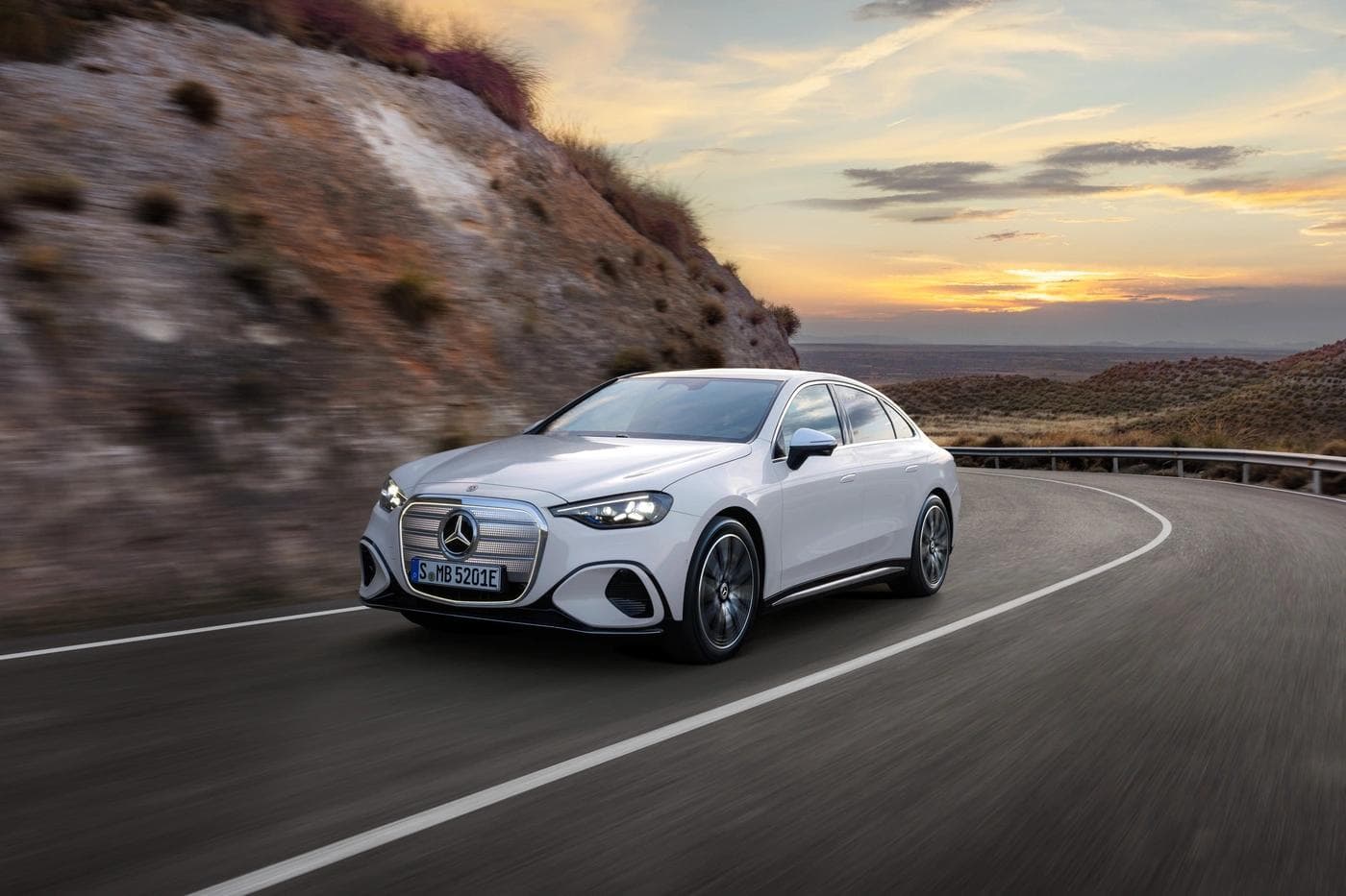 Mercedes-Benz C-Class with EQ Technology in white driving on a desert road at sunset, showing the illuminated grille and new design language.