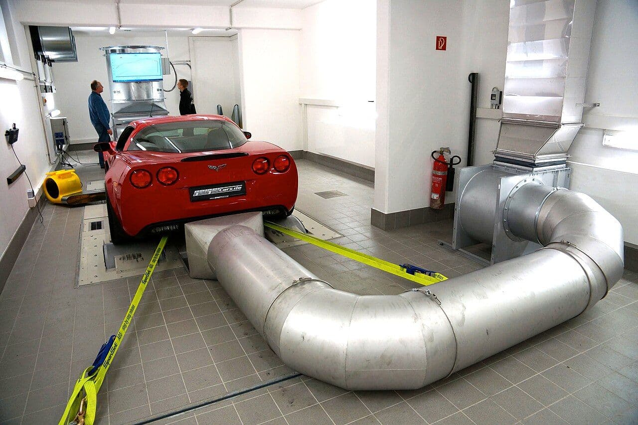 A red Chevrolet Corvette strapped onto a chassis dynamometer in a professional dyno testing room, with a large exhaust extraction system and two technicians observing data on a wall-mounted monitor.
