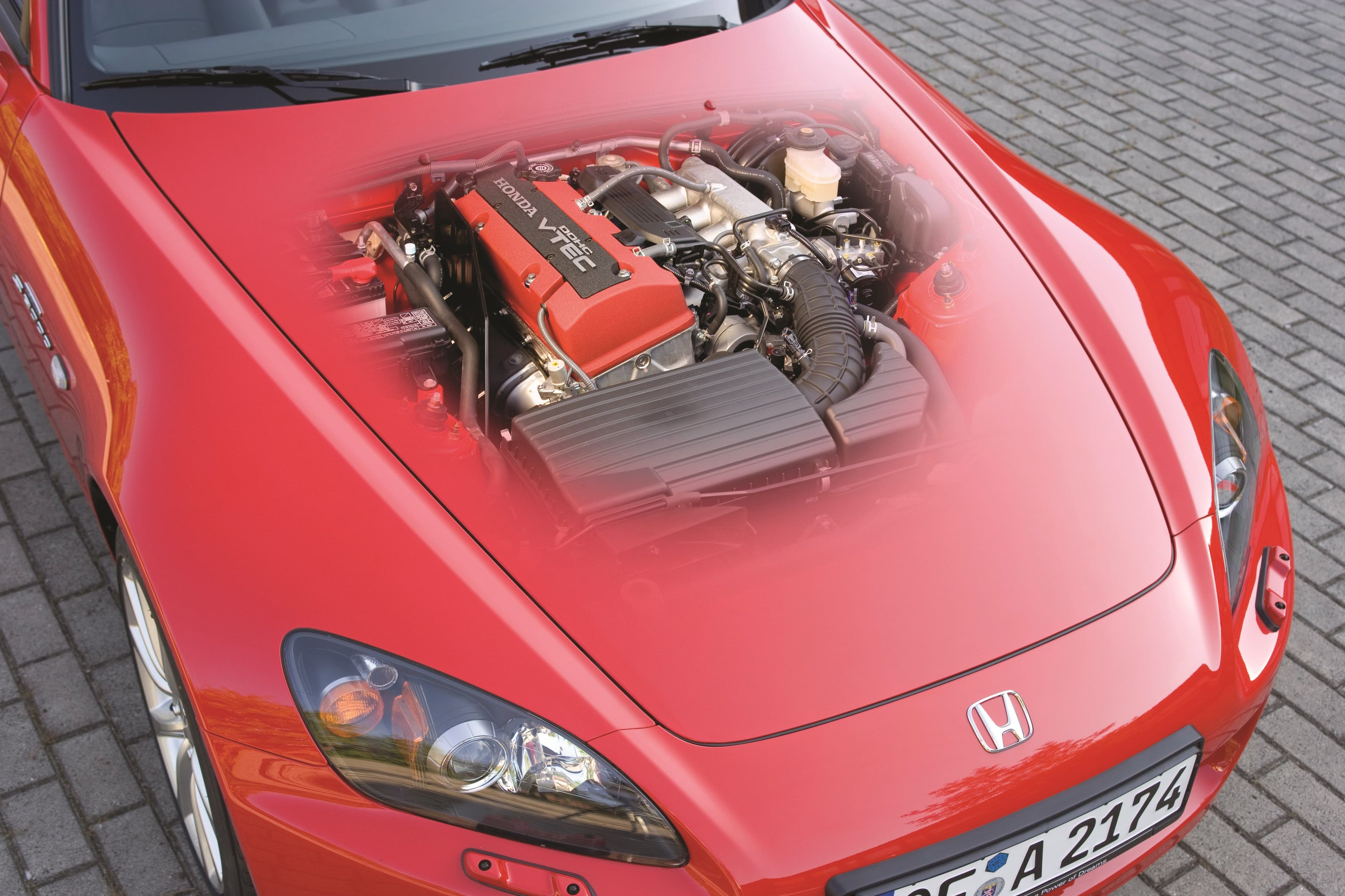 Close-up of the Honda S2000's F20C engine, featuring the iconic red valve cover and DOHC VTEC technology, partially visible through a transparent hood.