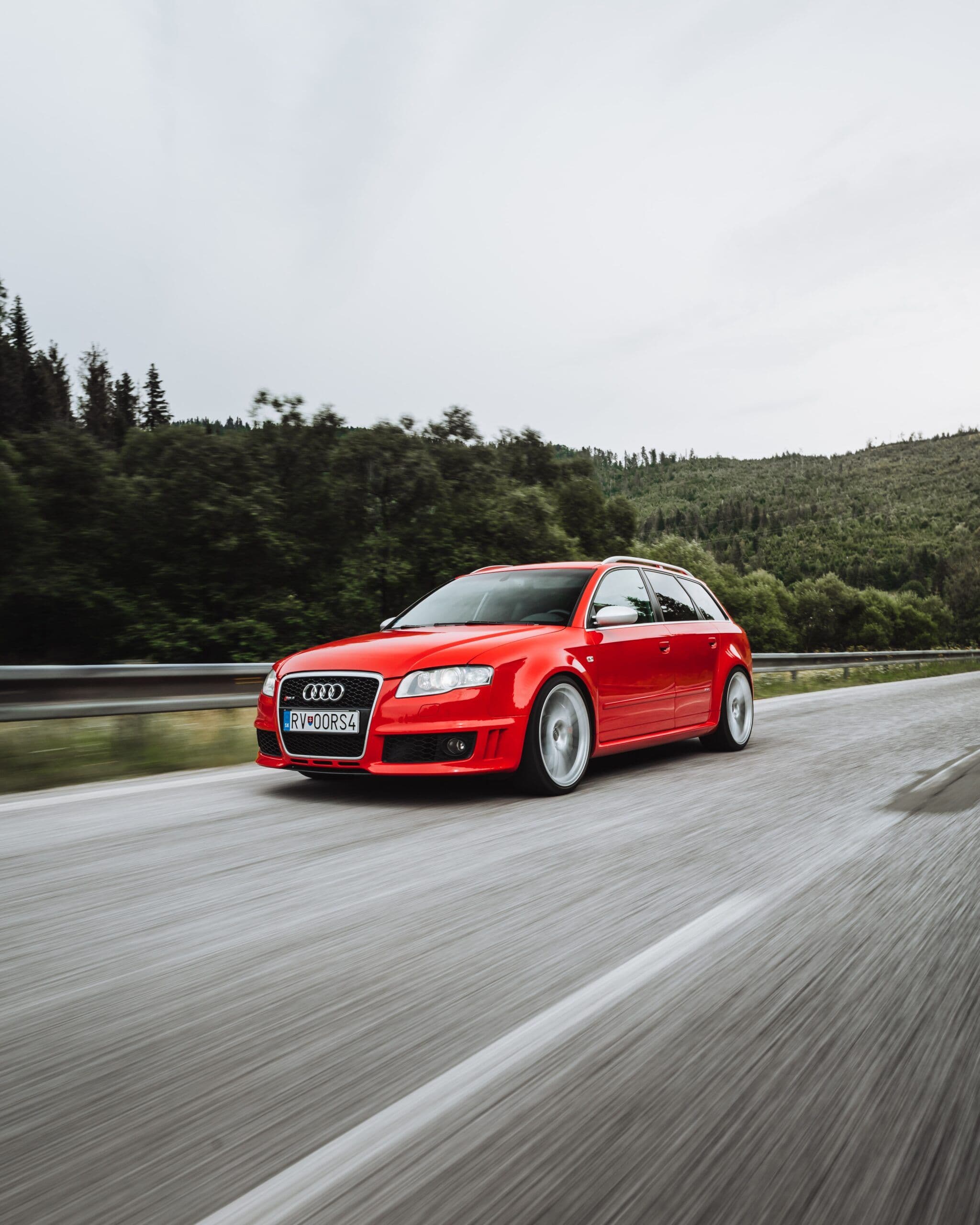 A red Audi RS4 Avant B7 driving at speed on a forested road, captured with motion blur to highlight its performance and dynamic stance.