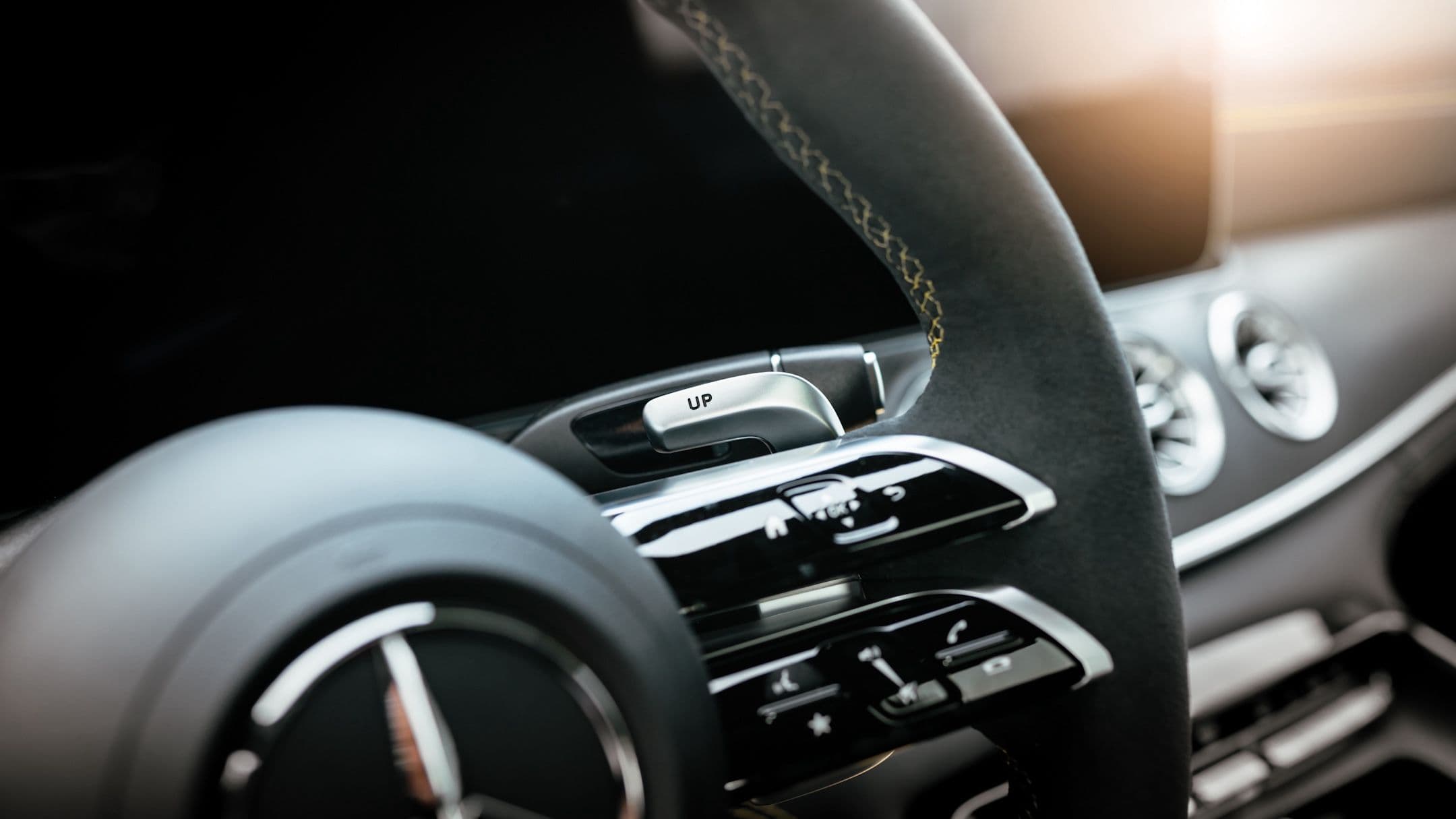 Mercedes-AMG paddle shifter marked 'UP' on a flat-bottom steering wheel with contrast stitching