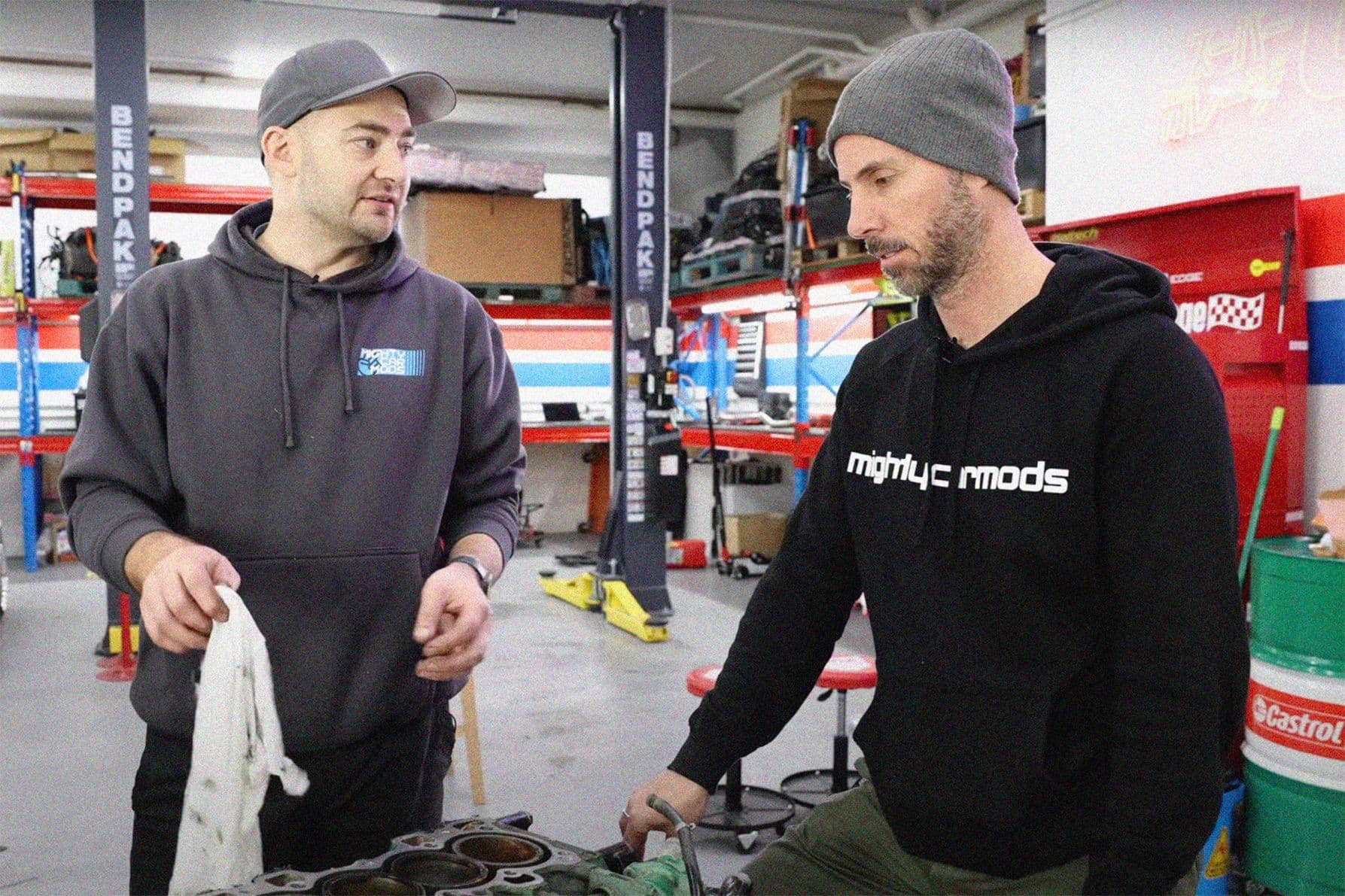 The hosts of Mighty Car Mods, Marty and Moog, working on an engine build inside their garage workshop, wearing branded hoodies surrounded by tools and automotive parts.