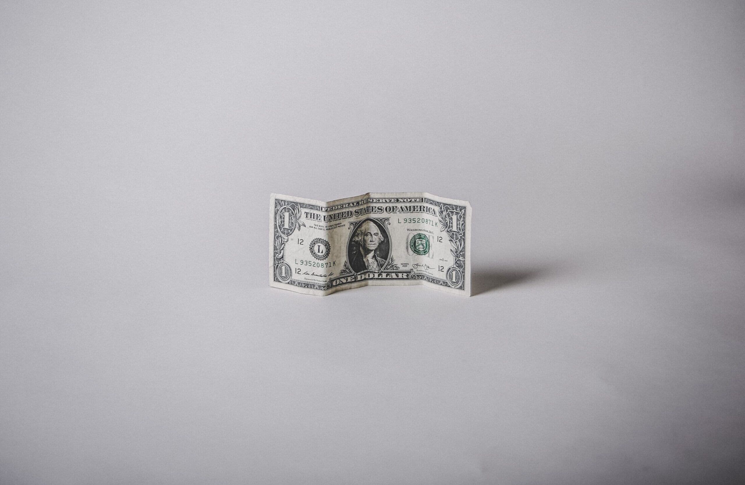 Crumpled US one-dollar bill standing upright on a minimalist grey background, symbolising low cost or minimal budget