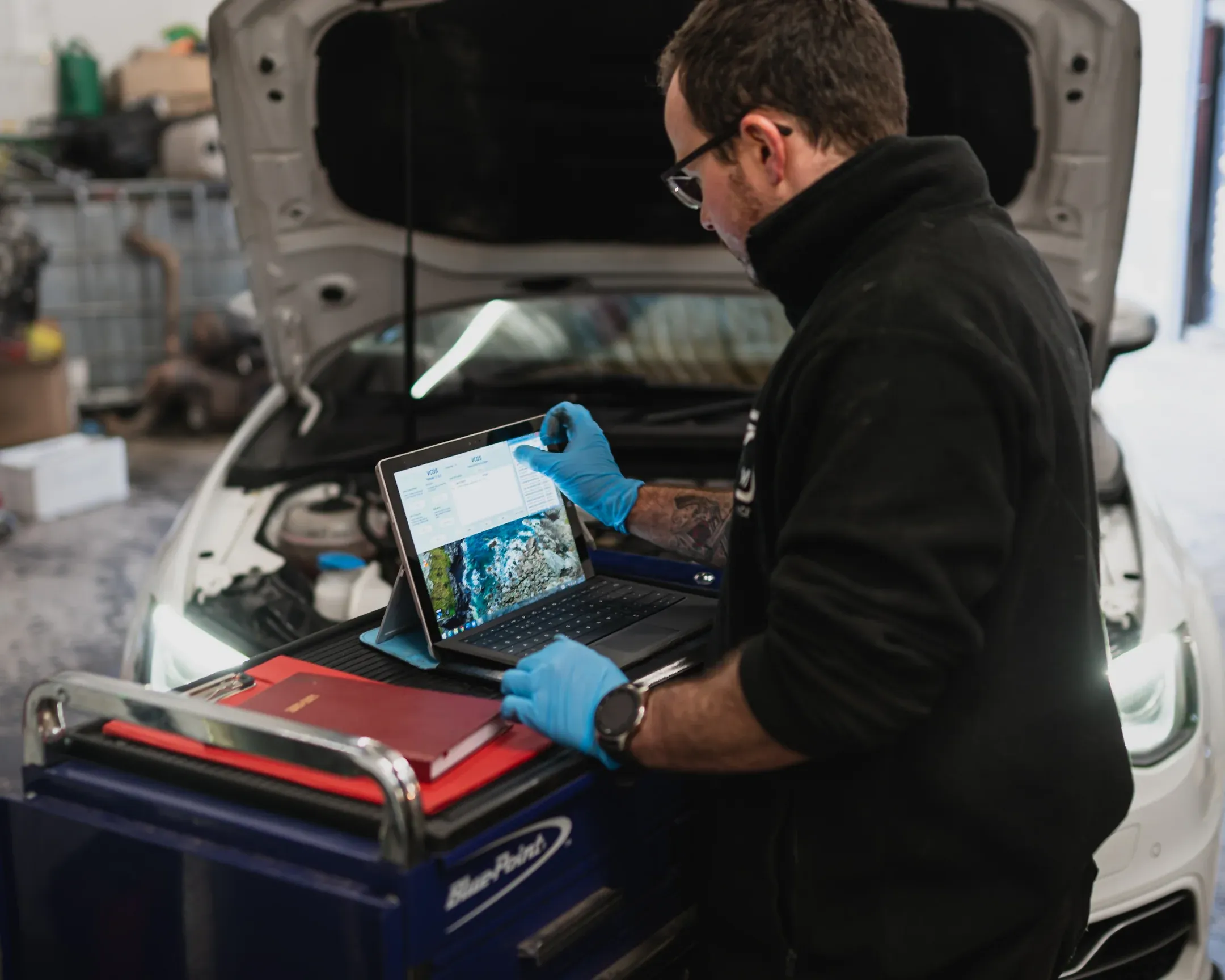 Automotive technician conducting a pre tuning diagnostic inspection using a laptop on a workshop tool cart, with a vehicle bonnet open in the background.