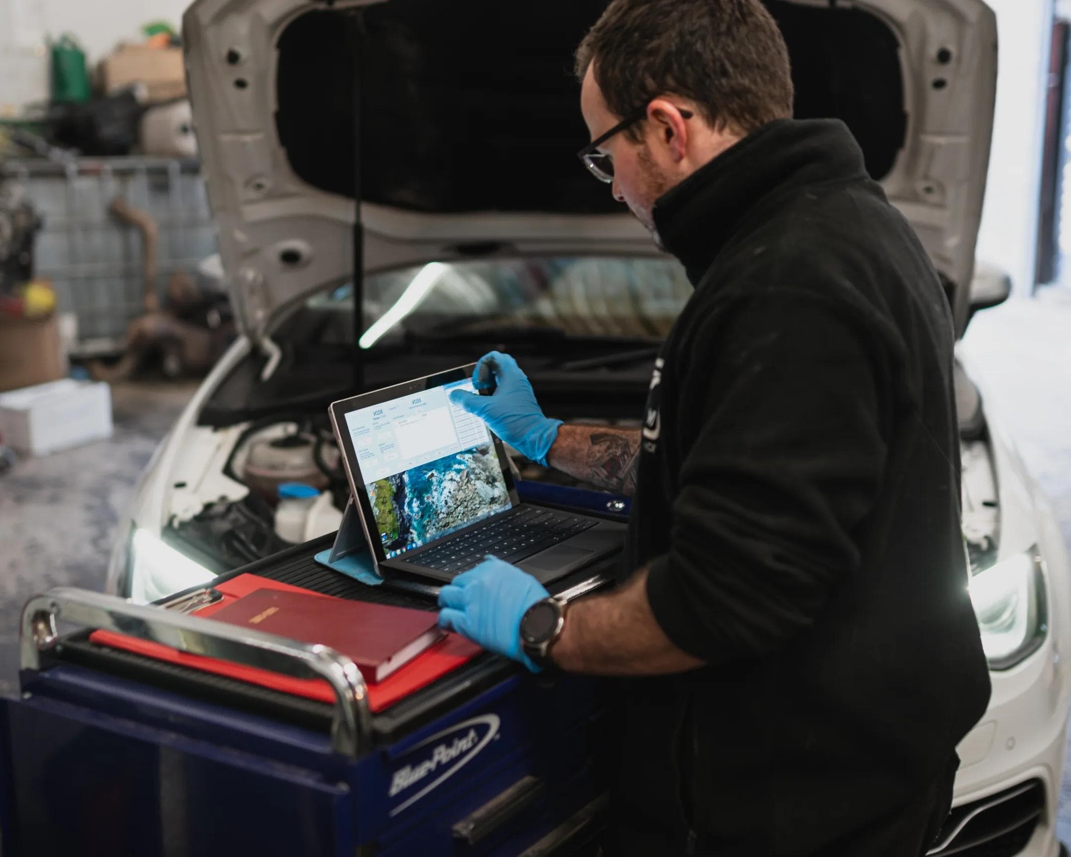 Automotive technician conducting a pre tuning diagnostic inspection using a laptop on a workshop tool cart, with a vehicle bonnet open in the background.