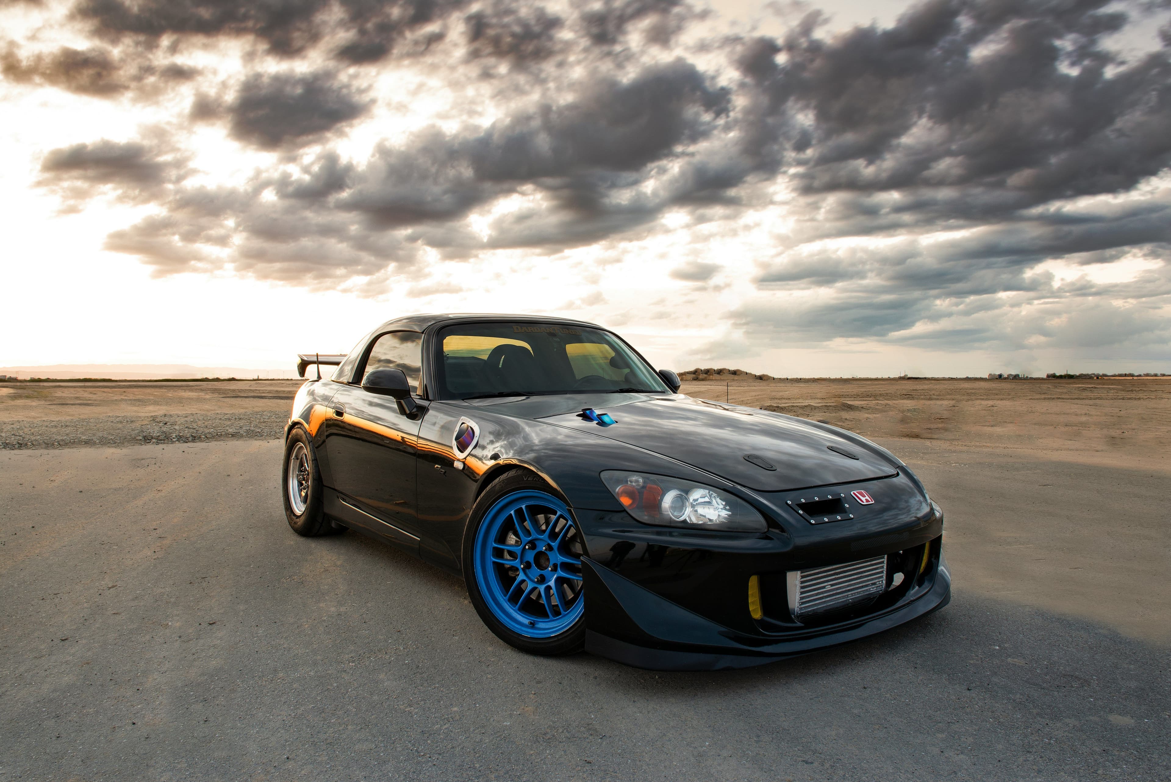 Black Honda S2000 with blue wheels and a racing setup, parked in a desert under dramatic clouds. Modified for drag racing with aerodynamic upgrades.