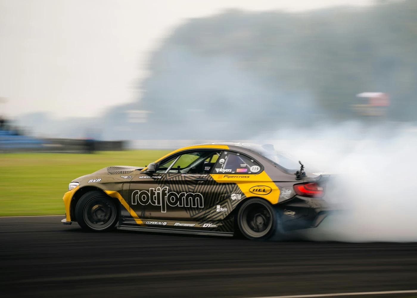 Jack Stapleton's Rotiform-sponsored BMW M2 drift car generating heavy tyre smoke during a competition run, number 119 visible on the door with Wheel Pros and Corbeau branding on the livery