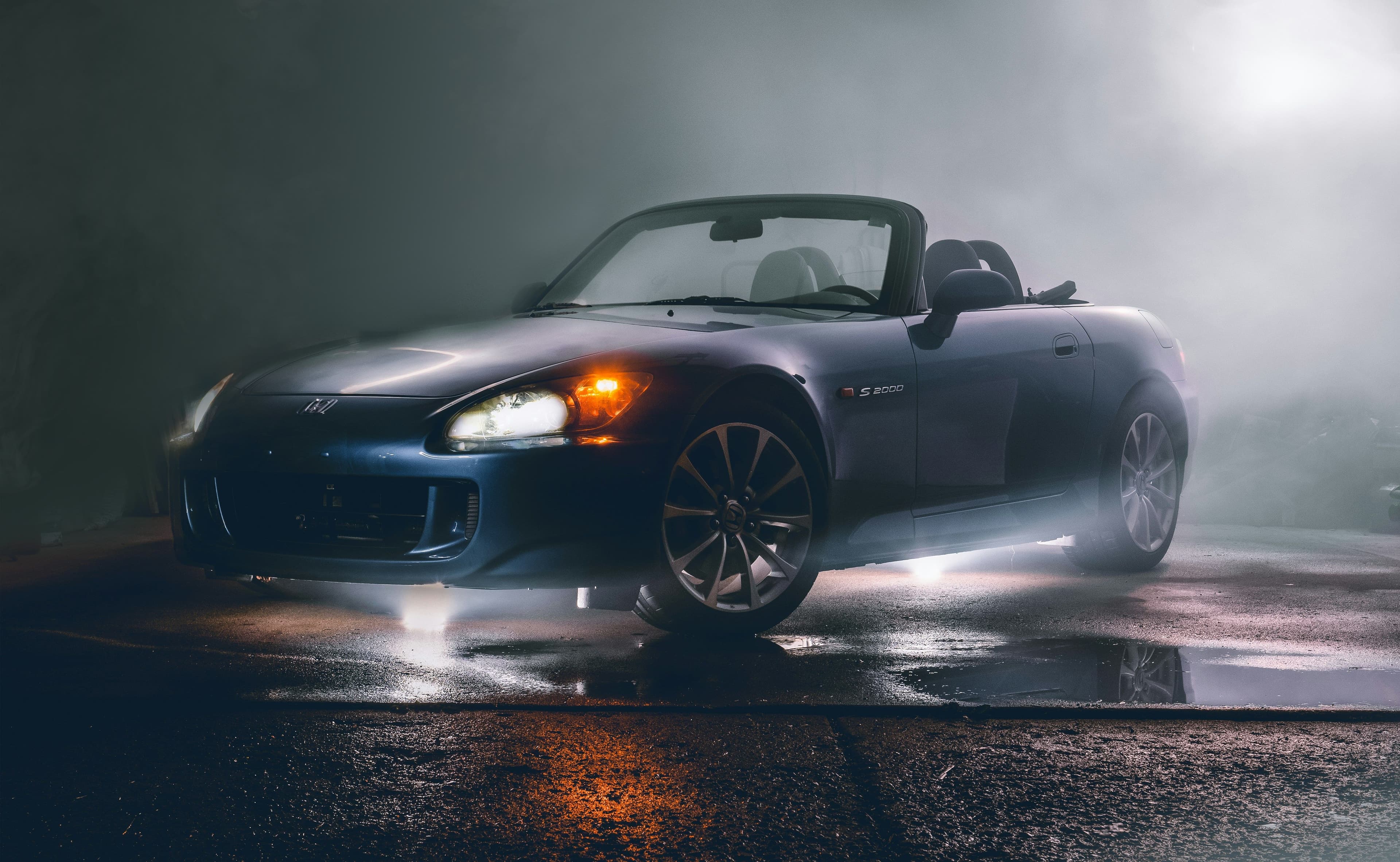 Blue Honda S2000 convertible with headlights on, illuminated in a misty parking lot. Moody lighting emphasizes its sleek design.
