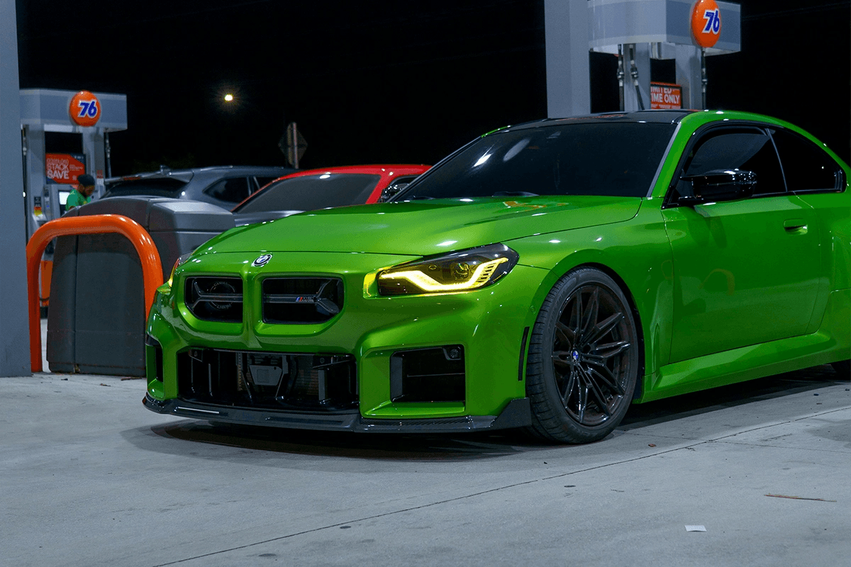 Green BMW G87 M2 parked at a petrol station at night, shown from a front three-quarter angle with headlights illuminated.