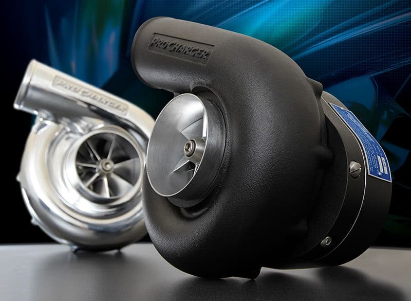 Two ProCharger centrifugal superchargers displayed on a table, with one unit finished in matte black and the other in polished chrome, both showcasing their precision impeller blades against a stylised automotive-themed backdrop.