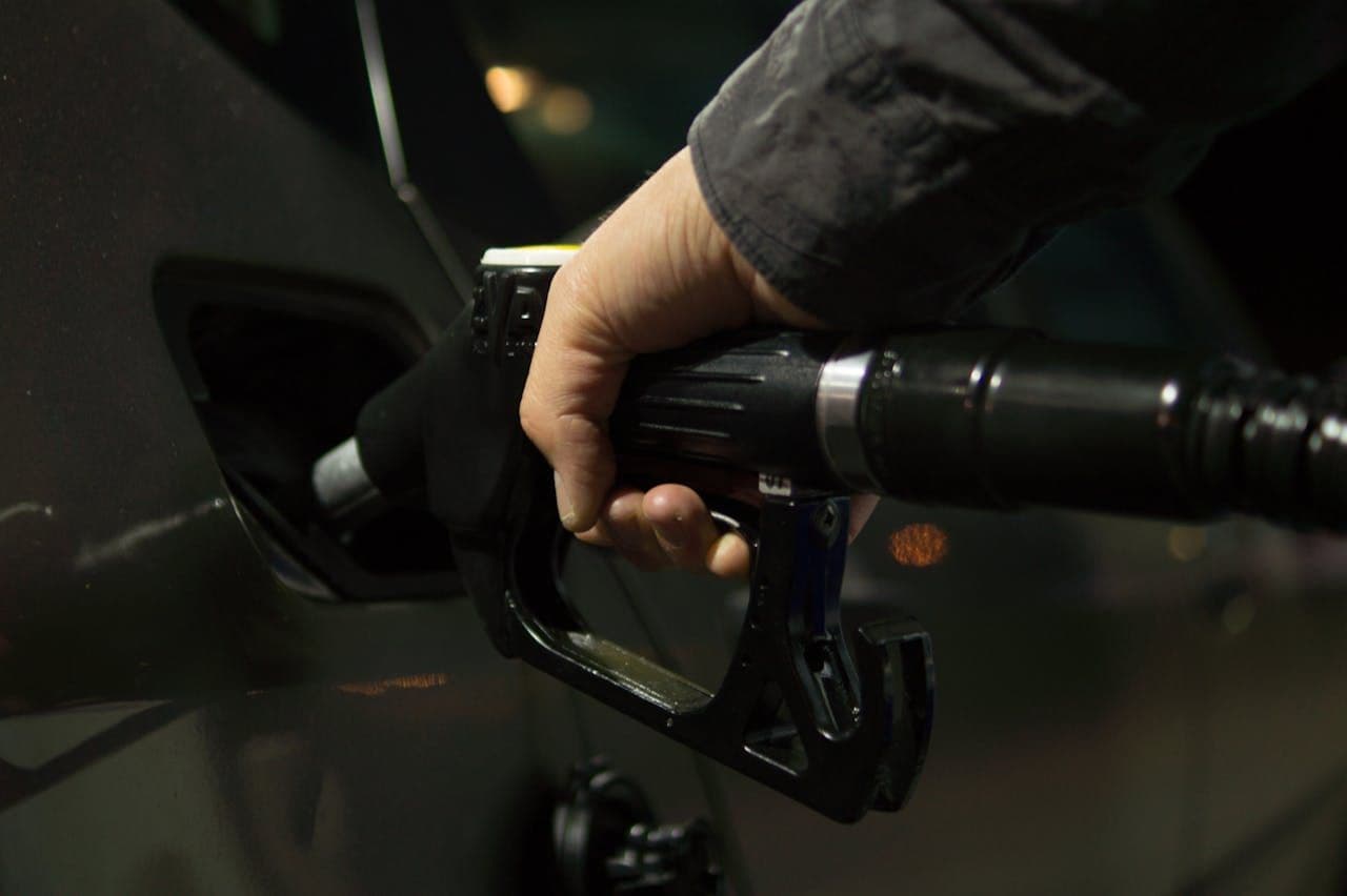 Close-up of a person refuelling a vehicle with a black fuel nozzle at night, with soft reflections on the car’s paint and blurred lights in the background.