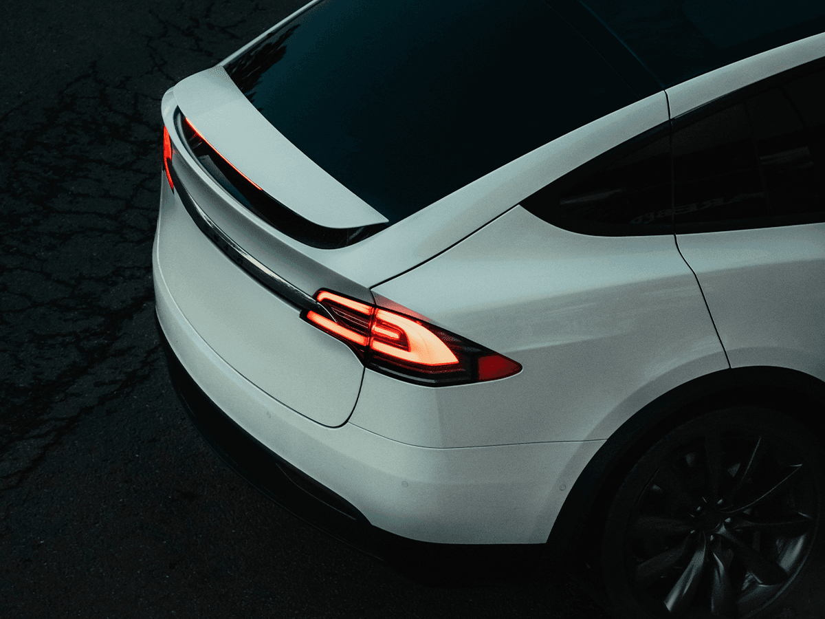 A white Tesla Model X captured from a high rear angle at dusk, showcasing its illuminated taillights, sleek aerodynamic design, and black multi-spoke wheels. The car’s rear spoiler and tinted windows add to its futuristic aesthetic.