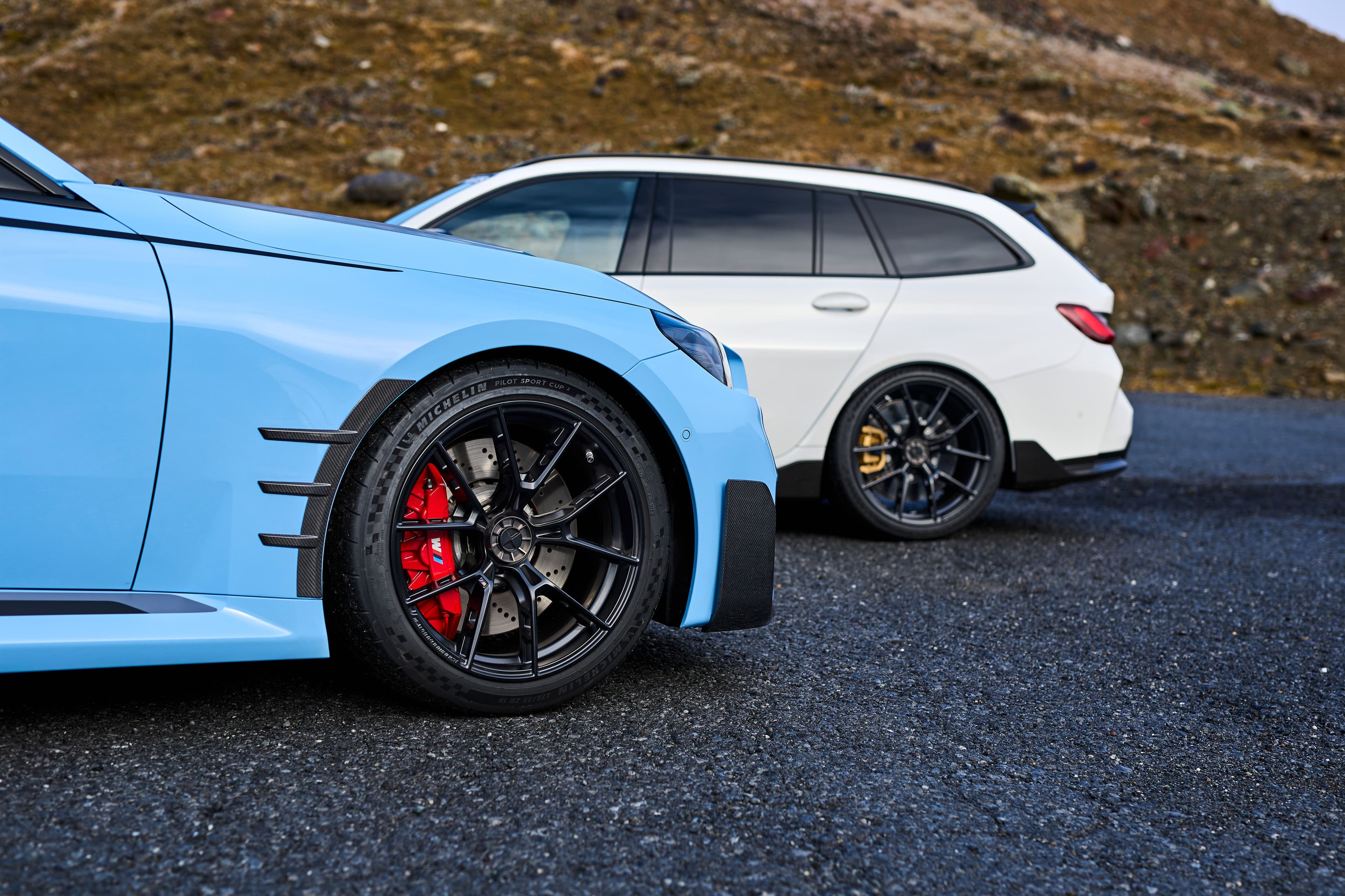 A blue BMW M2 and a white BMW M3 Touring parked on a mountain road, both equipped with the new BMW center-lock wheels. The M2 features red brake calipers, while the M3 Touring has gold calipers.