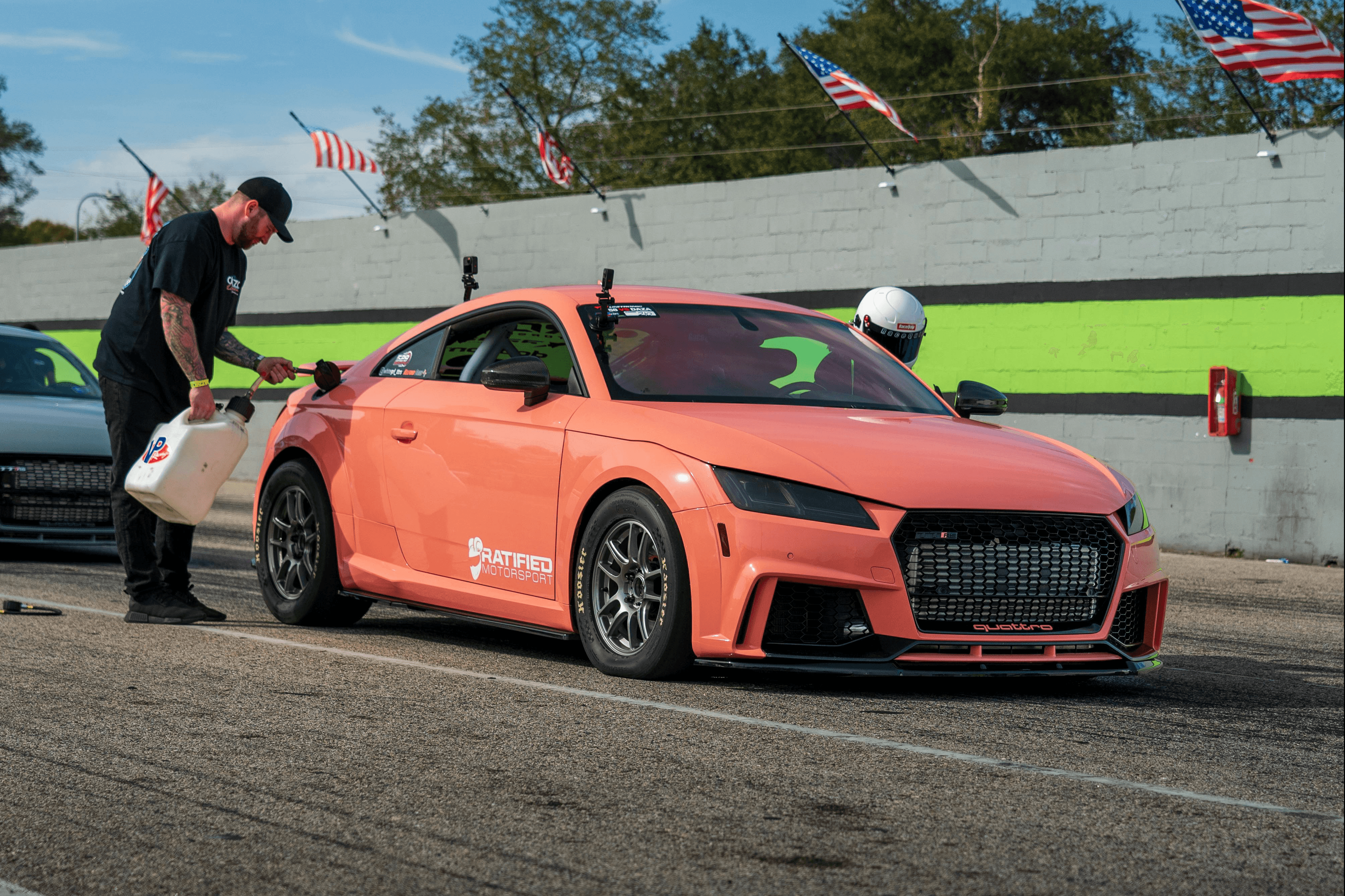 A coral-colored Audi TT RS race car is parked in the pit lane while a crew member refuels it with a jerry can. The driver, wearing a white helmet, is ready inside the car, with American flags waving in the background.