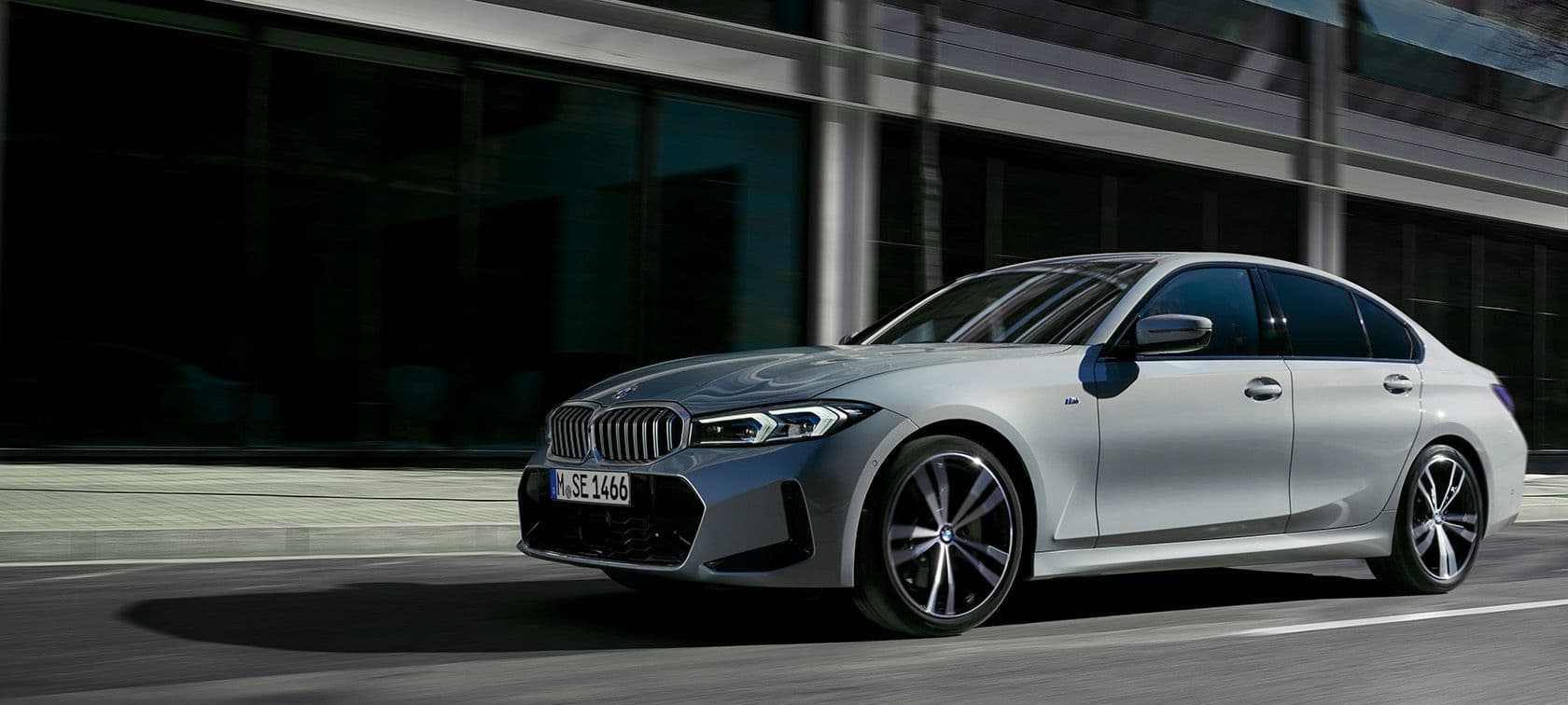 A silver BMW 3 Series saloon driving through a city street with modern glass architecture in the background, captured mid-motion in a dynamic side profile.