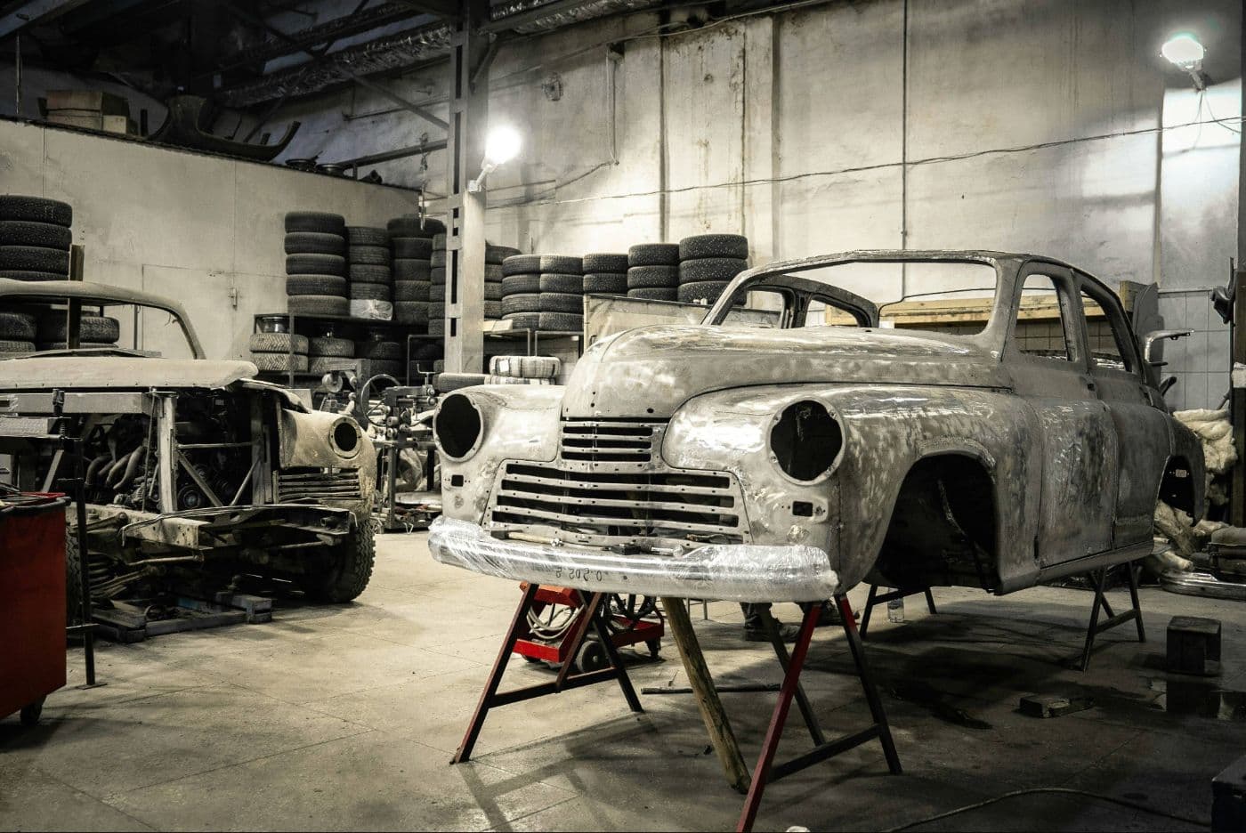 Bare metal classic car shells undergoing restoration in a workshop, with spare tires stacked in the background.