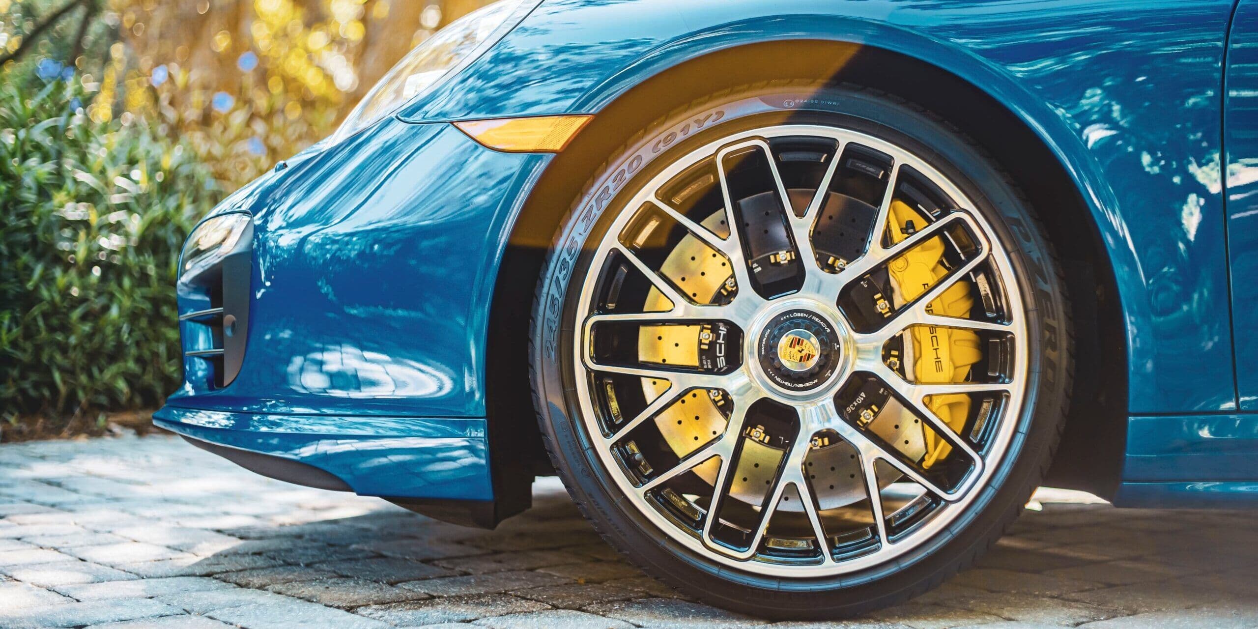 Close-up of a Porsche sports car’s front wheel with a yellow brake caliper and multi-spoke alloy rim, parked on cobblestone pavement with natural greenery in the background.